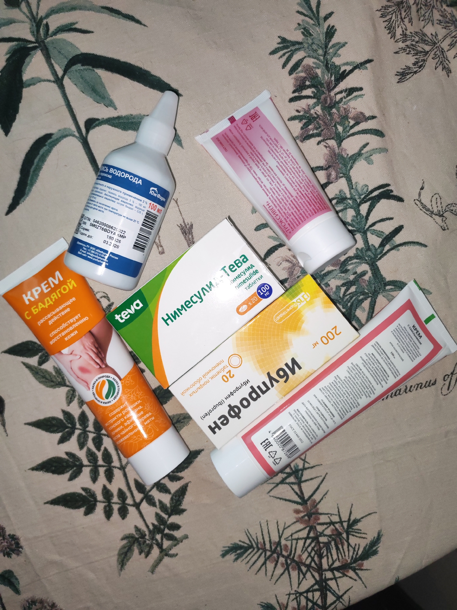 The image shows a collection of over-the-counter medications on a floral-patterned surface. Among them are:
- A bottle of "Hydrogen Peroxide 3%", made by "Yuzh Farm," with the volume of 100 ml, manufactured in Russia.
- "Krem s Bodyagoy" (Cream with Bodyaga), intended for skin recovery.
- A package of "Nimesulide-Teva" tablets (20 tablets of 100 mg each).
- A box of "Ibuprofen" 200 mg tablets, made by Pharmstandard.
- Two tubes of cream: one is "Troxiven" gel and the other is a "Cream with Comfrey" containing plant extracts, oils, and chondroitin.
The location in the city of Izhevsk, Russia is not explicitly mentioned in the text. However, one manufacturer address is listed as being in Kirov, Russia. There are no individuals visible, and the environment appears to be indoors.