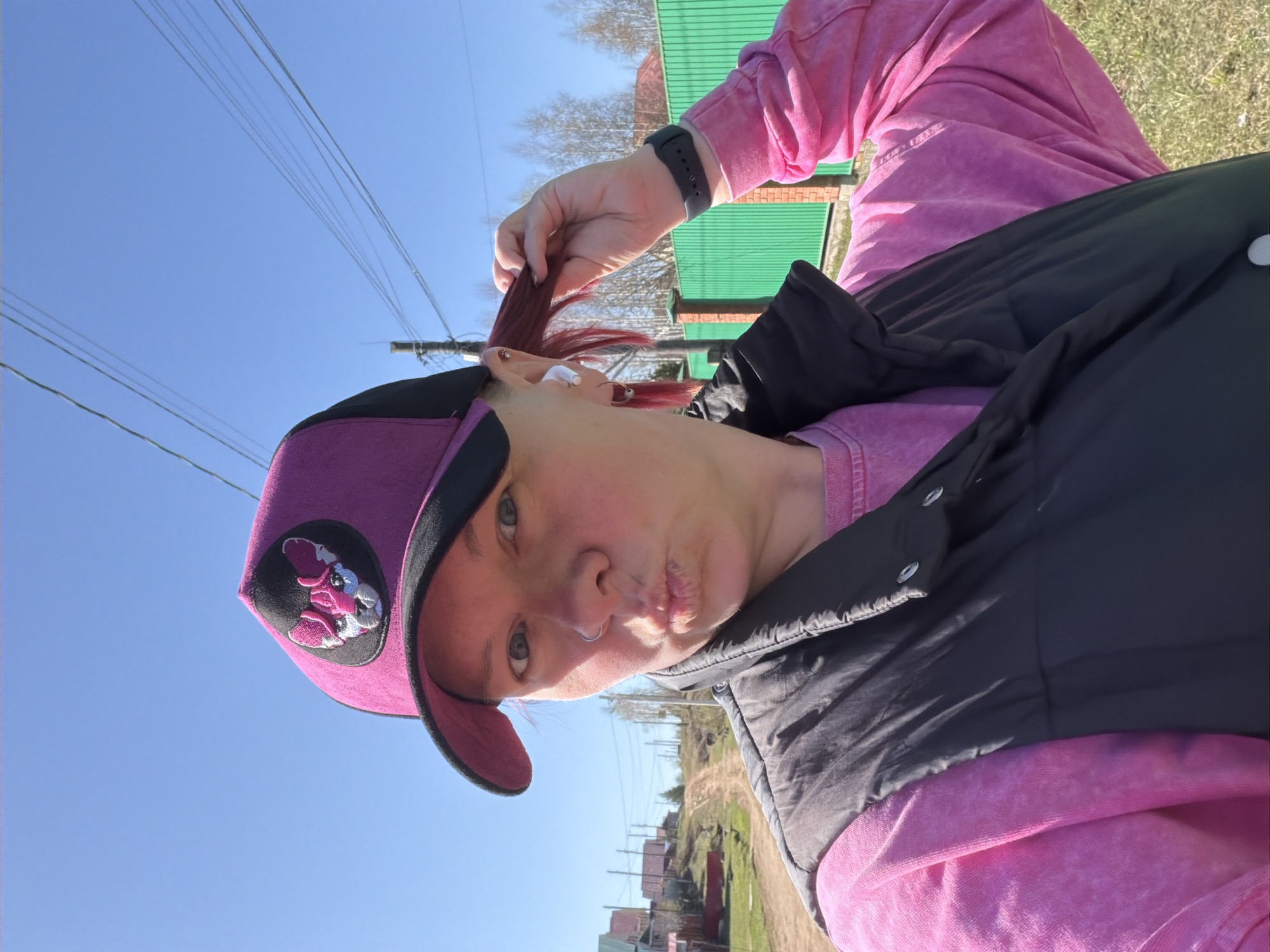 A person with red hair is visible from the chest up, wearing a pink sweatshirt, a black vest, and a purple cap with an embroidered design. The person is holding a strand of their hair near their ear, and their lips are pursed in a kissing gesture. The sky above is clear and blue. In the background, there is a row of small buildings, a dirt road, utility poles with wires, and green vegetation. The location is Chaikovsky, Russia.