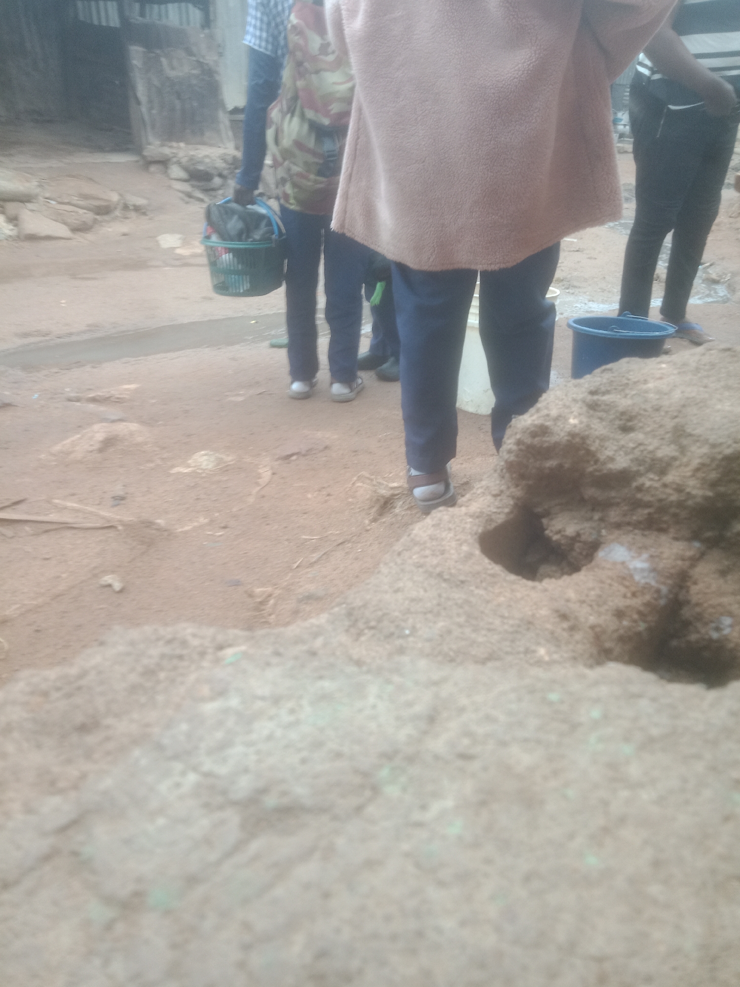 The image depicts a ground-level view of several individuals in what appears to be an outdoor, possibly urban or semi-urban, setting. The ground is predominantly dirt and sand, with some larger rocky or earthy formations in the foreground and background. In the mid-ground, a person is visible from the waist down, wearing dark pants and a light-colored, possibly fleece, top. To their left, another individual is seen from the knees down, wearing dark pants and a backpack with a camouflage pattern, carrying a green plastic basket filled with dark items. Further to the left, another person's legs are visible, wearing dark pants and a backpack. In the background, more legs and lower torsos of people are visible, suggesting a group activity. There are at least two buckets present, one blue and one white, on the ground. The overall lighting suggests an overcast or shaded environment.