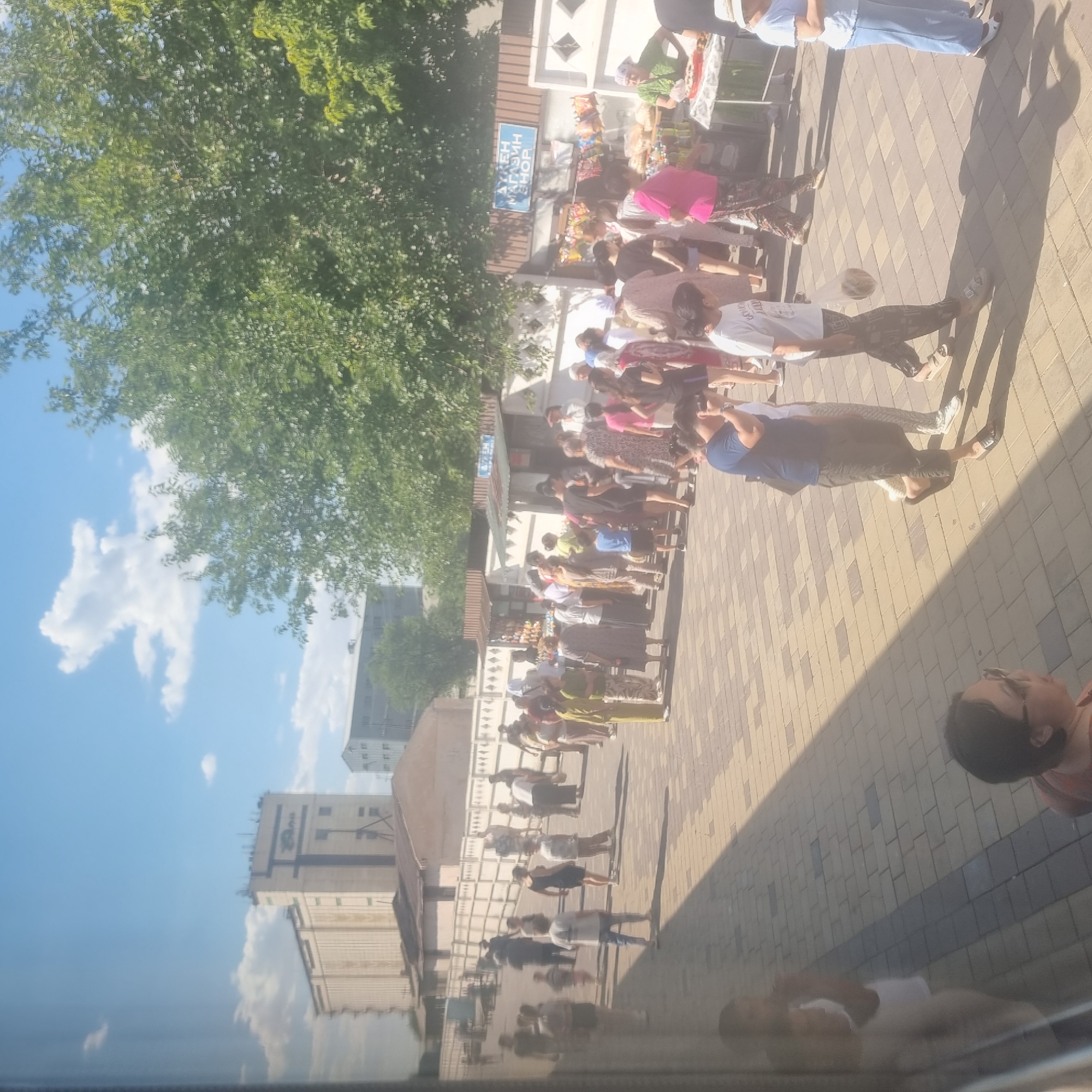 The image shows a busy street scene with a crowd of people gathered near stalls. The location is identified as the city of Turkistan, Kazakhstan. Several buildings are visible in the background, including a large industrial-looking structure on the left and smaller shops or market stalls in the middle ground. The ground is paved with interlocking bricks.
The people in the image are dressed in various casual clothing, with some individuals wearing headscarves. Many people appear to be walking or standing around the stalls, suggesting a market or a gathering place. Some of the stalls are displaying goods, such as food items and drinks. The sky is blue with scattered clouds, and trees are visible on the right side of the image, indicating an outdoor setting on a sunny day. Reflections are visible on the left side of the image, suggesting the photo was taken through a window.