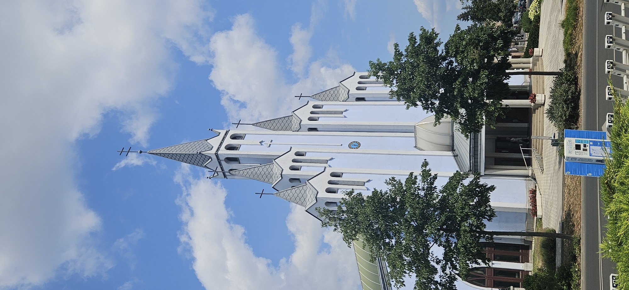 The image shows the facade of a light blue and white church with several spires and crosses under a partly cloudy blue sky. The architecture features tall, narrow windows and decorative cornices. In the foreground, there are two green trees on either side of the church entrance. A paved walkway leads up to the entrance, which has a ramp and steps. To the right of the entrance, a circular clock face is visible on the church wall. In the lower foreground, a blue information kiosk is situated near a road. The scene appears to be in the city of Hévíz, Hungary, given the presence of the church which is recognizable as the Church of the Holy Spirit in Hévíz.