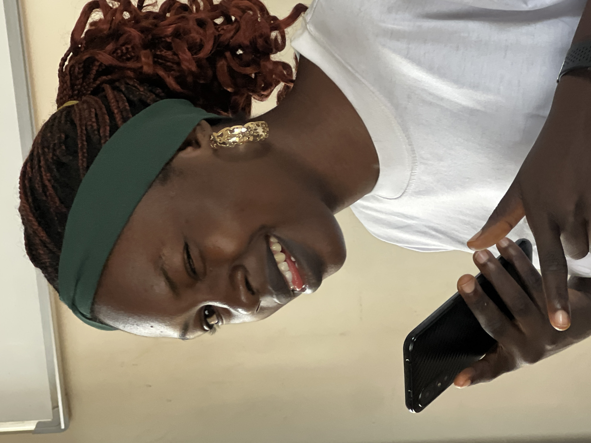 A medium shot captures a young Black woman from the chest up, looking to her left and winking. She is wearing a green headband, large gold hoop earrings, and a white t-shirt. Her dark brown hair is braided and pulled back into a ponytail. She holds a black smartphone in her left hand and her right index finger is extended, possibly interacting with the screen. Her mouth is slightly open, revealing her tongue and teeth, and she appears to be smiling. The background consists of a beige wall and a white board with a grey frame. The scene suggests a casual setting, possibly in an office or classroom in Jos, Nigeria.