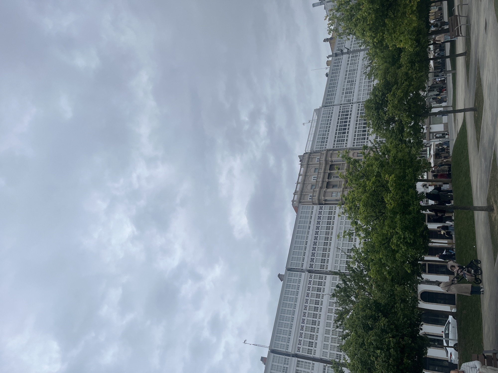 The image captures a street view in A Coruña, Spain, under a cloudy sky. A prominent building featuring rows of white-framed windows dominates the background. A line of trees runs along the right side of the frame, partially obscuring the building and sidewalk. People are walking along the sidewalk. A woman in a tan coat pushes a stroller.