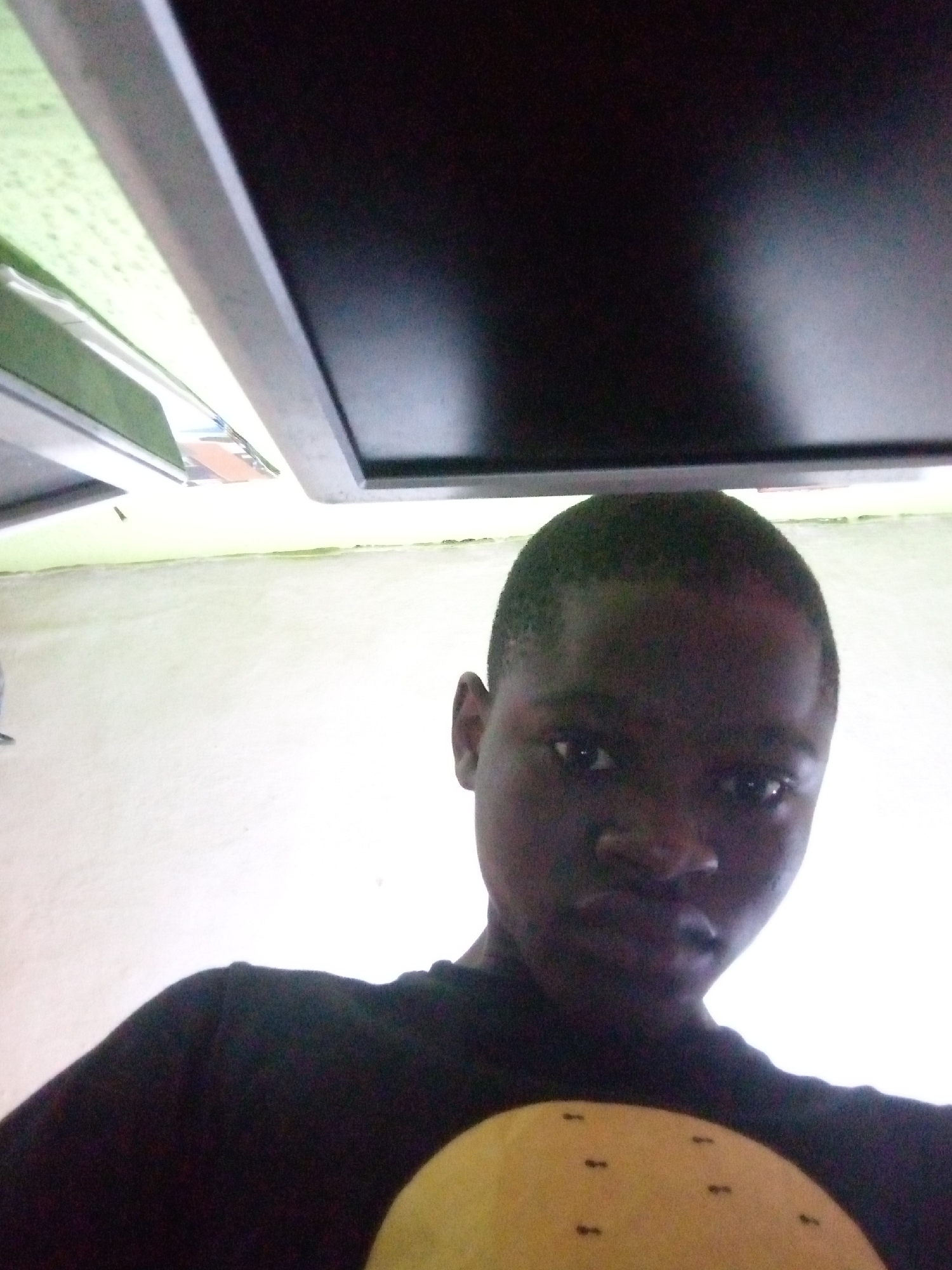The image shows a close-up, head-and-shoulders shot of a young Black male. He is looking directly at the camera. He has dark skin, short dark hair, and appears to be wearing a black t-shirt. Part of a yellow object with small black markings is visible in the lower right corner of the frame. The background is largely out of focus, but appears to be a light-colored wall. Above the man's head, a portion of a dark, rectangular object, possibly a monitor or a ceiling fixture, is visible. The lighting is bright, casting some glare on the man's face and the background. The image is oriented sideways. No specific location in Shangisha, Nigeria can be identified from the visual information provided.
