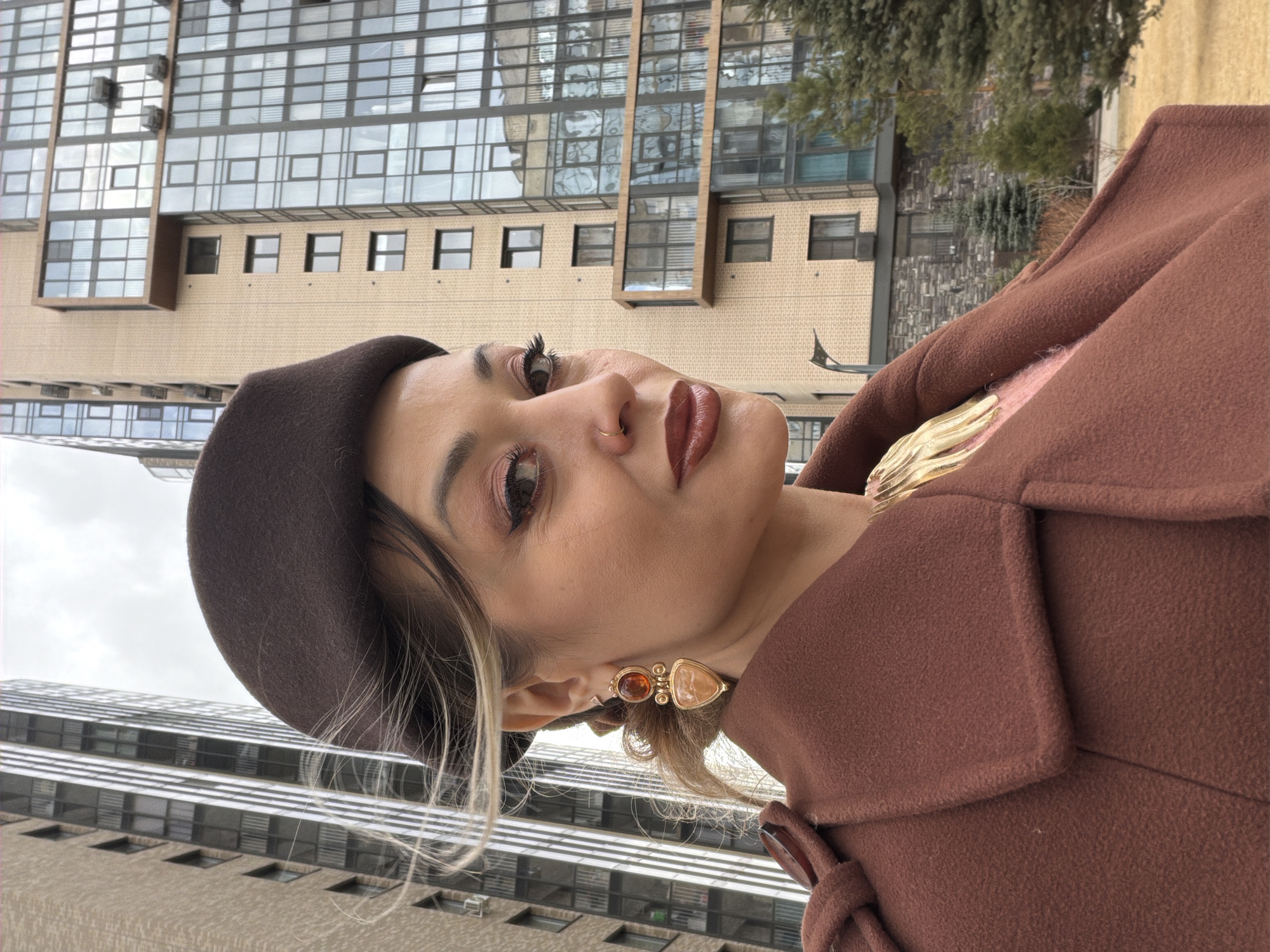 A woman is standing in front of two buildings in what could be the city of Krasnoyarsk, Russia, based on architectural style. The woman is wearing a brown beret, a brown coat with a pink top underneath, and earrings with a golden setting and brown and tan stones. She has light brown hair with blonde highlights, and she is wearing makeup. In the background, there is a building with a reflective glass exterior and another building with a beige brick exterior. There is some vegetation visible. The photograph is taken from a low angle, looking up at the woman.