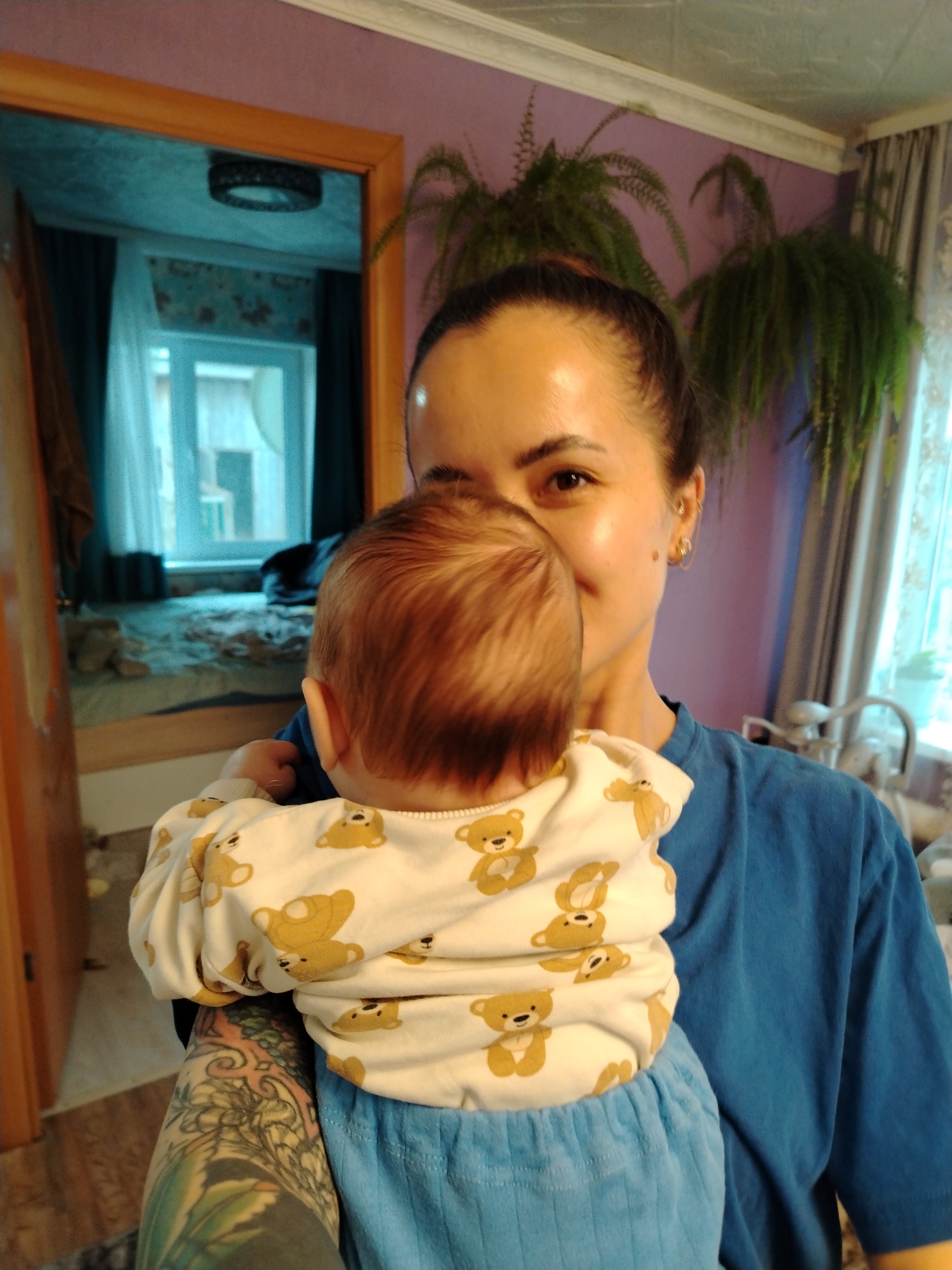 A woman holds a baby in a room with lavender walls and ferns hanging from the ceiling. The woman has dark hair pulled back in a bun and wears a blue t-shirt. The baby wears a white onesie with teddy bears and blue pants. The woman has a tattoo on her left arm. A doorway reflects a room with a bed and a window. The location is Гожан, Russia.
