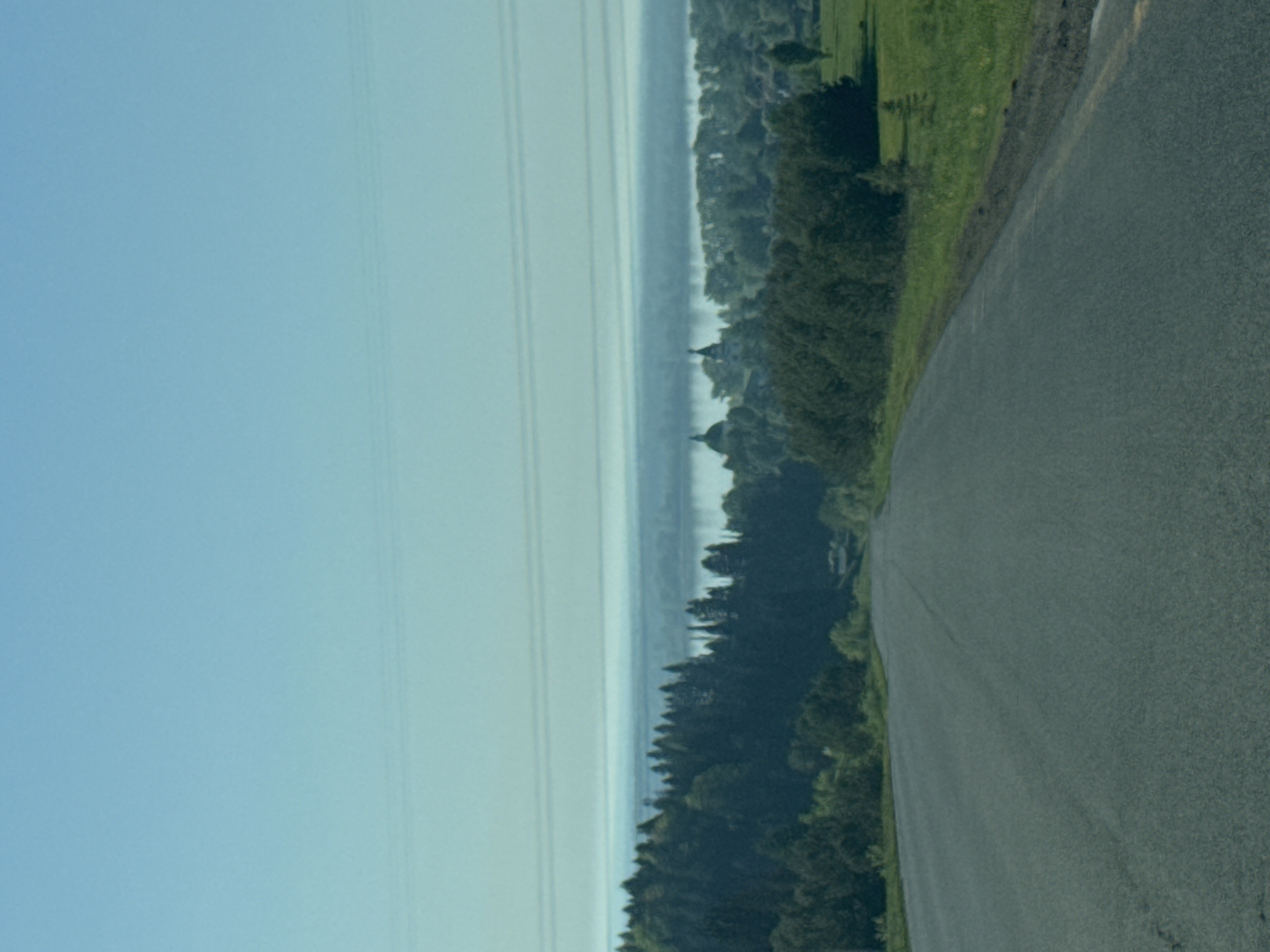 A landscape view of a road curving to the right. To the left of the road, trees and grass grow. Further to the left, a lake or river is visible, and power lines run along the left side of the image. The sky is light blue. The location is Makarovo, Russia.