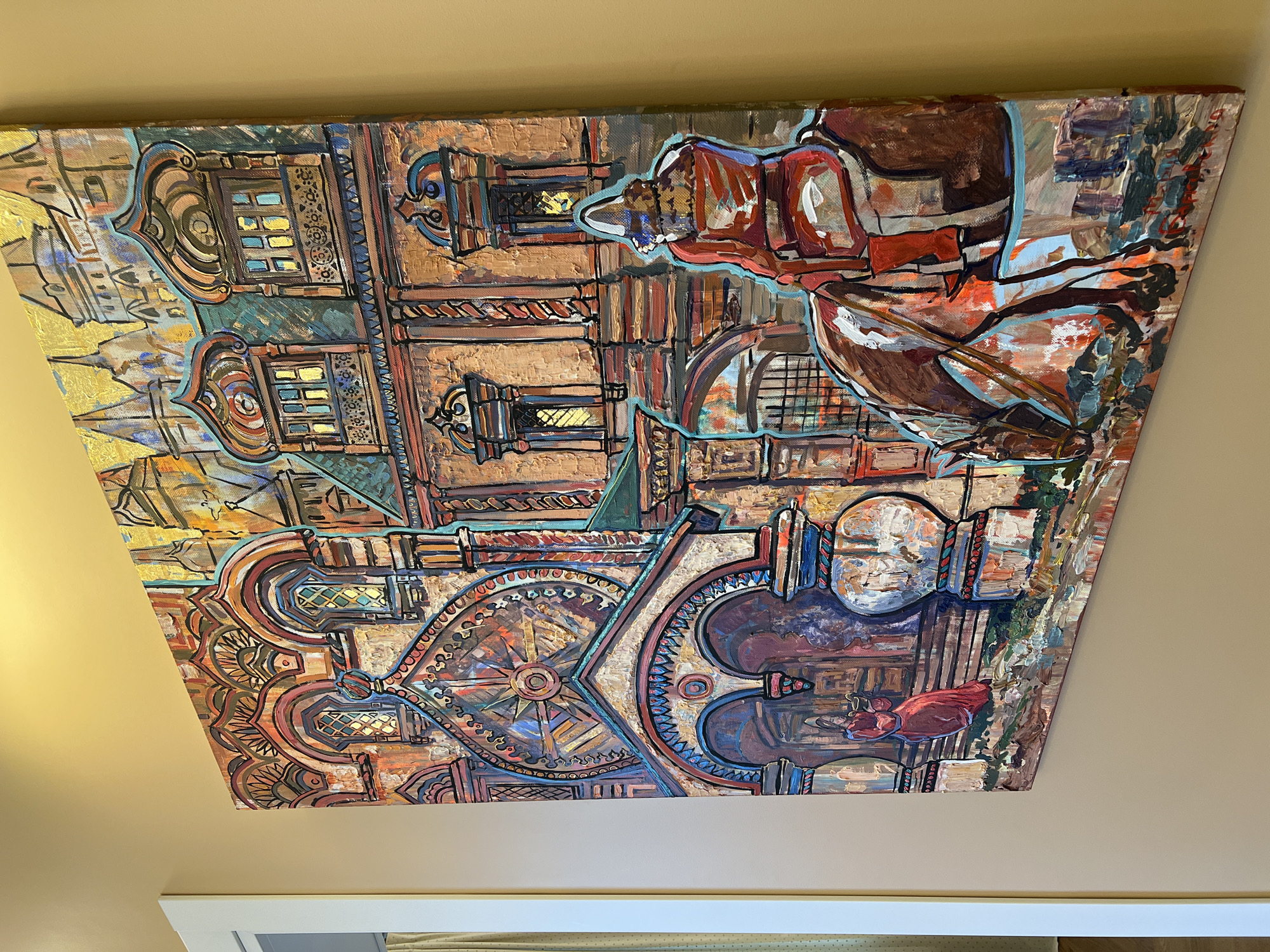 The image is a close-up, angled shot of a vibrantly colored, textured painting mounted on a light beige wall. The painting depicts a stylized architectural scene reminiscent of St. Basil's Cathedral in Moscow, Russia, with its onion domes and ornate facades rendered in bold brushstrokes of reds, oranges, blues, and golds. In the foreground, a figure on horseback is shown from behind, facing away from the viewer. The rider is wearing a red garment, and the horse is a rich brown. To the left, a woman in a red dress stands in an arched entryway of the building. The painting is richly detailed with intricate patterns and decorative elements, suggesting a historical or religious setting. The painting is unsigned, but its style is suggestive of Russian folk art or avant-garde interpretations of traditional Russian architecture. There is no indication of the specific city of Yakunino, Russia, in the image.