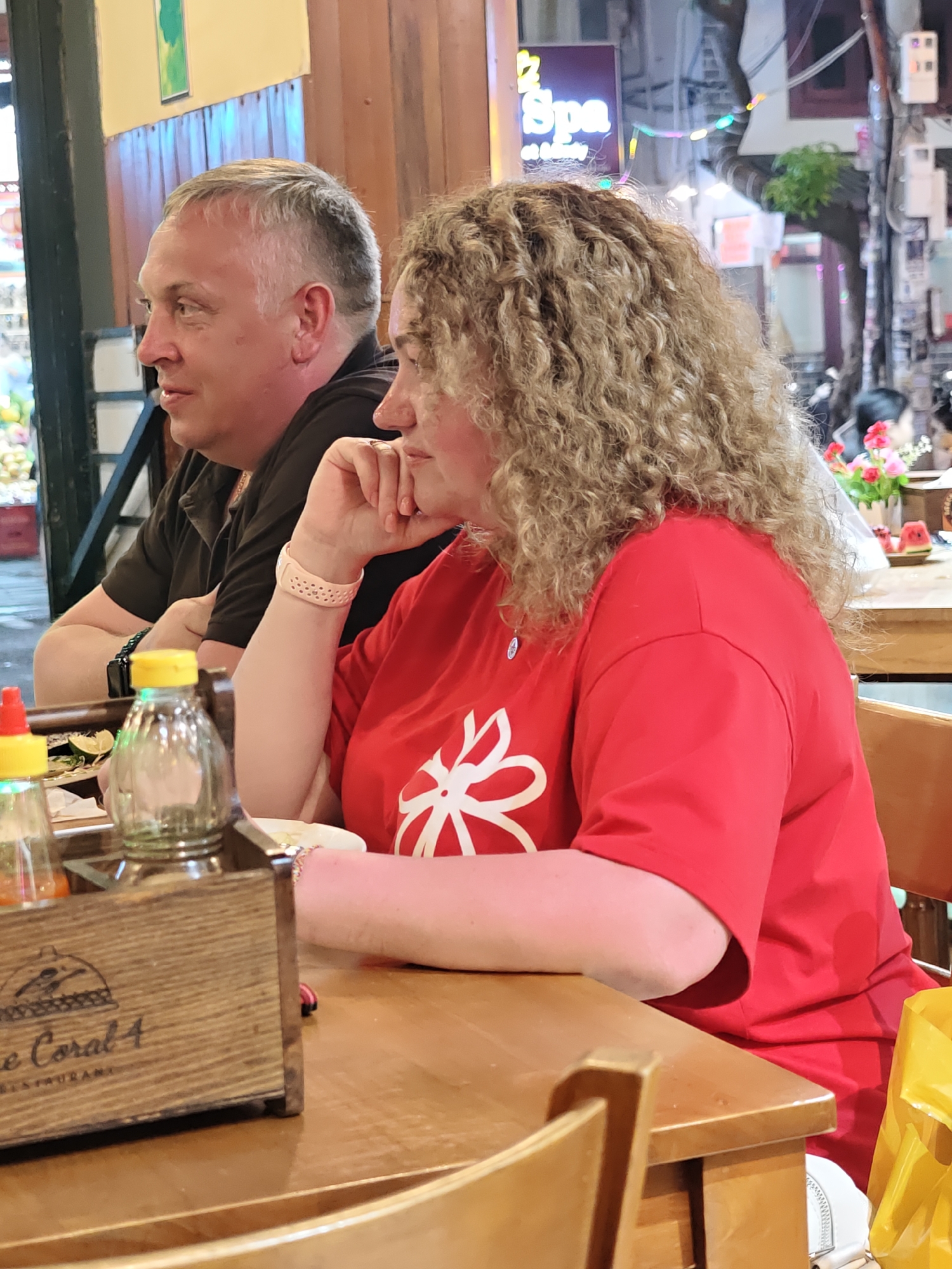 Two people are seated at a wooden table in what appears to be an outdoor restaurant setting. The man on the left is wearing a dark polo shirt and has short gray hair. He is looking to his left, with his mouth slightly open as if speaking. The woman on the right has curly blonde hair and is wearing a bright red t-shirt with a white floral design on the chest. She is resting her chin on her hand, looking to her right. On the table in front of them are two glass bottles with yellow and red caps, and a wooden container with some text on it. In the background, there are signs and lights, suggesting it is evening or nighttime. The setting appears to be a busy street in Vietnam, possibly Nha Trang Ward, as indicated by the location context. The overall mood seems relaxed, with the couple enjoying a meal or drinks together. The visible text on the wooden container includes "The Coral 4 RESTAURANT".