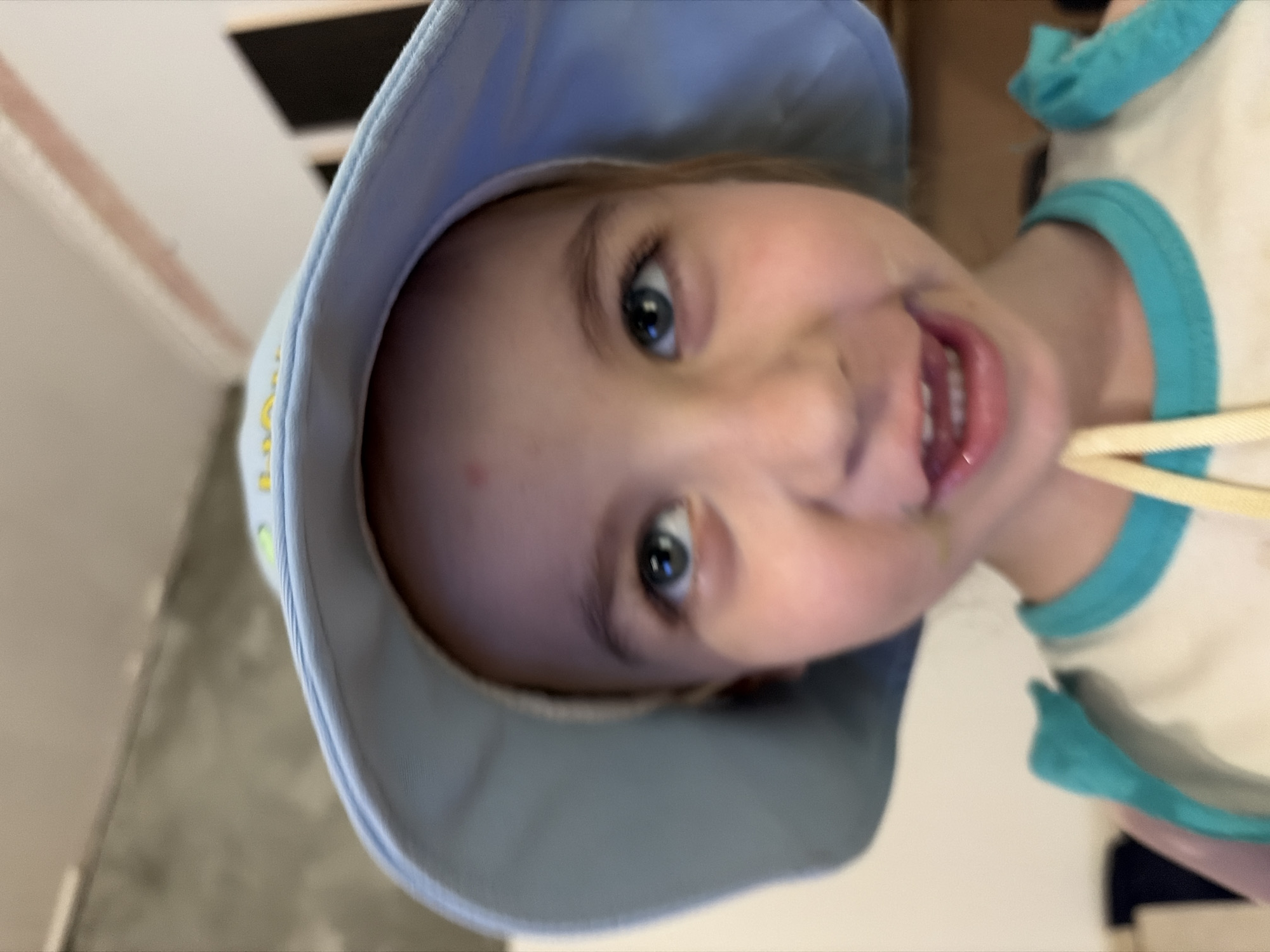 The image, taken from a low angle, features a young girl wearing a light blue sun hat and a white tank top with turquoise trim. She is looking upwards and to her right with a wide, open-mouthed smile. The background is blurry, but appears to be an indoor setting with neutral-colored walls and possibly some dark objects visible. No specific landmarks or features indicating the city of Tambov, Russia, are discernible in the image.