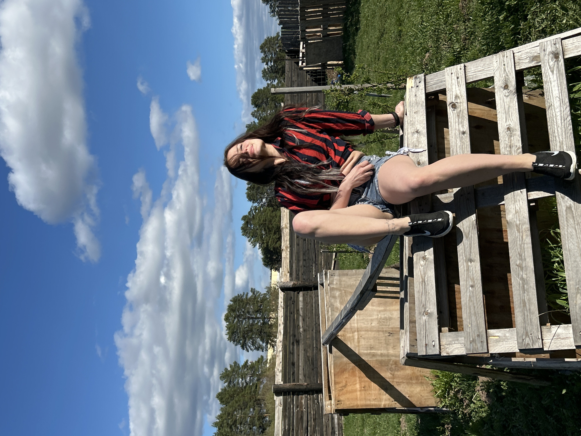 The image shows a woman sitting on a wooden structure, possibly a pallet or raised platform, in an outdoor setting. She is wearing a red and black striped long-sleeved shirt and denim shorts, and black and white athletic shoes. Her hair is long and dark. She is squinting and her head is tilted back as if she is looking toward the sky. The background includes a partly cloudy blue sky, greenery, and wooden fencing or construction. The location appears to be rural or on the outskirts of a built environment. There is no information to confirm if the location is Rusalevka, Russia.