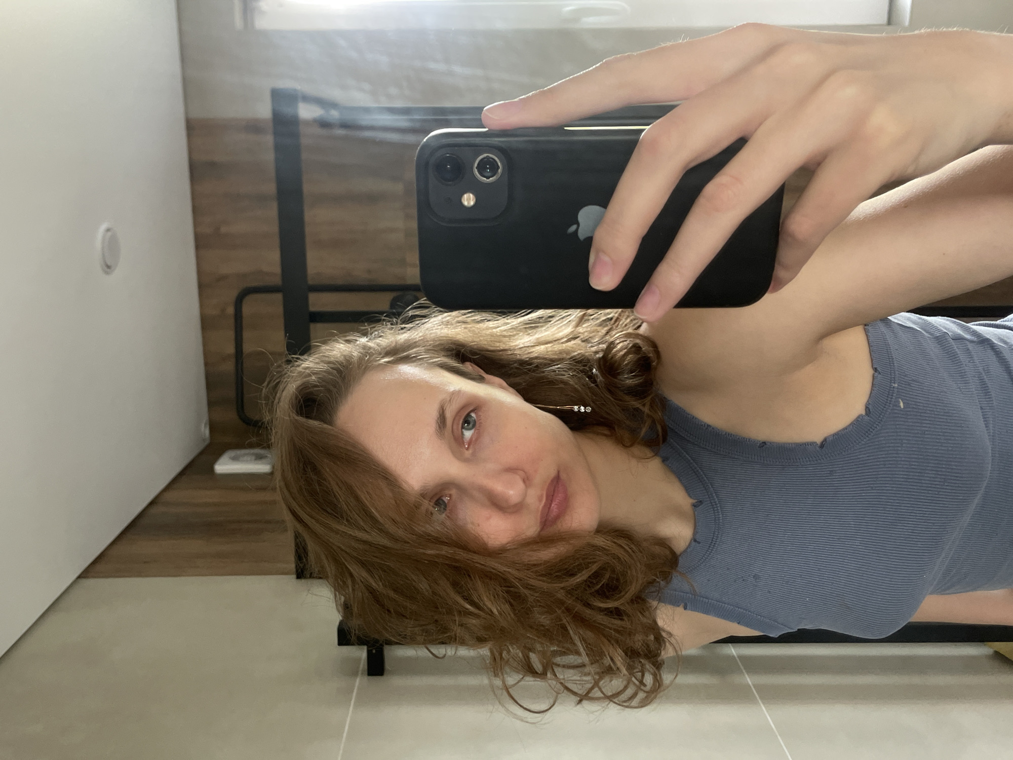 A woman is taking a selfie in what appears to be a bathroom. She is holding a black iPhone with two rear cameras. She has light brown hair and is wearing a ribbed blue tank top. The background includes a white door with a circular fixture, a wooden paneled wall, light beige floor tiles, and part of a metal bed frame.