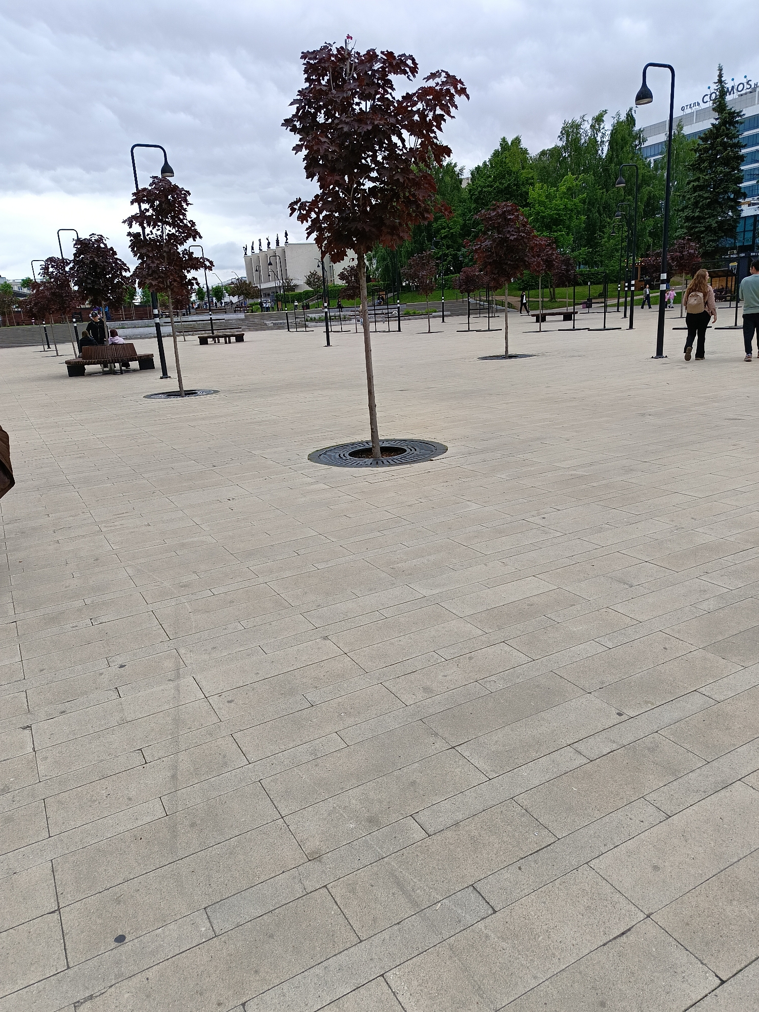 The image displays an urban pedestrian plaza in Izhevsk, Russia, under an overcast sky. The ground is paved with rectangular light grey concrete or stone slabs, laid in a running bond pattern. Several young trees with dark reddish-brown foliage are planted at regular intervals across the plaza, each featuring a circular black metal grate at its base. Numerous black, modern streetlights with distinctive curved heads are interspersed among the trees.
On the left side of the plaza, two individuals are seated on a dark wooden bench with metal supports. One person wears a dark jacket and a cap, while the other wears a lighter-colored garment. In the right midground, two individuals walk away from the camera. The leading person, a woman, wears a light-colored top, dark trousers, and a light-colored backpack. To her left, a man wears a light green top and dark trousers.
In the background on the left, a light-colored building with a prominent roofline adorned with several human-like statues is visible; this structure is identifiable as the Udmurt State Philharmonic Hall. To the right of the Philharmonic, a tall, multi-story modern building with a glass facade is visible, displaying the words "ОТЕЛЬ СОМОS" and "COSMOS" on its upper section, identifying it as the Cosmos Hotel Izhevsk. Further in the background, densely packed green trees and other urban structures define the broader city environment.