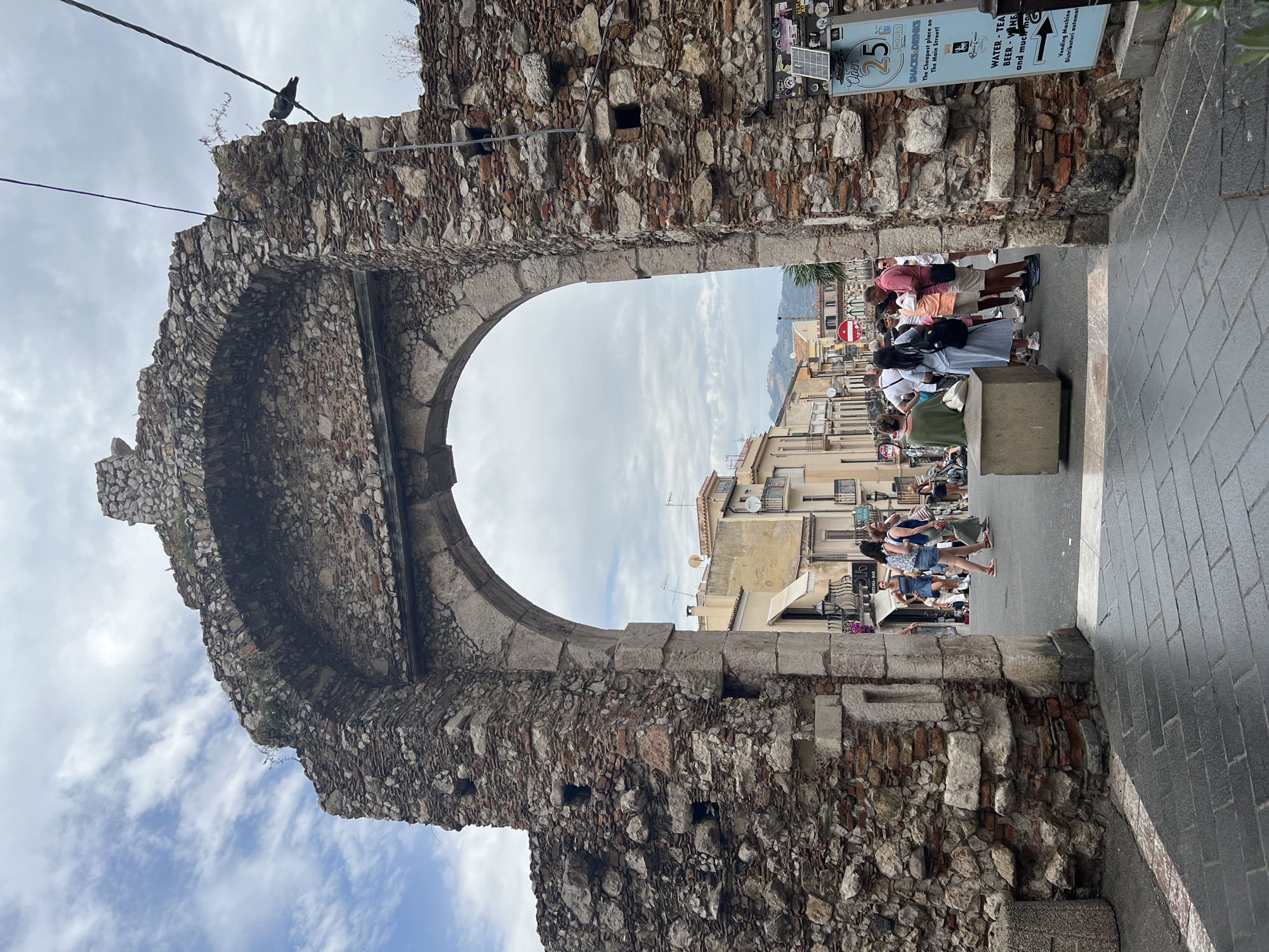 The image captures a view through a large, ancient stone archway, likely part of the Porta Messina in Taormina, Italy. The arch is constructed of rough-hewn stones and bricks, showing signs of age and decay, with some vegetation growing from cracks. Beyond the arch, a street lined with buildings, typical of Italian architecture with balconies and tiled roofs, is visible. The sky above is cloudy, with patches of blue. A group of people are gathered in the area beyond the arch, some standing and conversing, others walking. One woman sits on a concrete cube. A sign is attached to the stone structure on the right, advertising "Open 25 HOURS SNACKS & DRINKS" and "WATER - TEA - BEER - WINE and much mo...". A vending machine is also depicted below the sign. The foreground shows a paved walkway with a patterned stone surface.