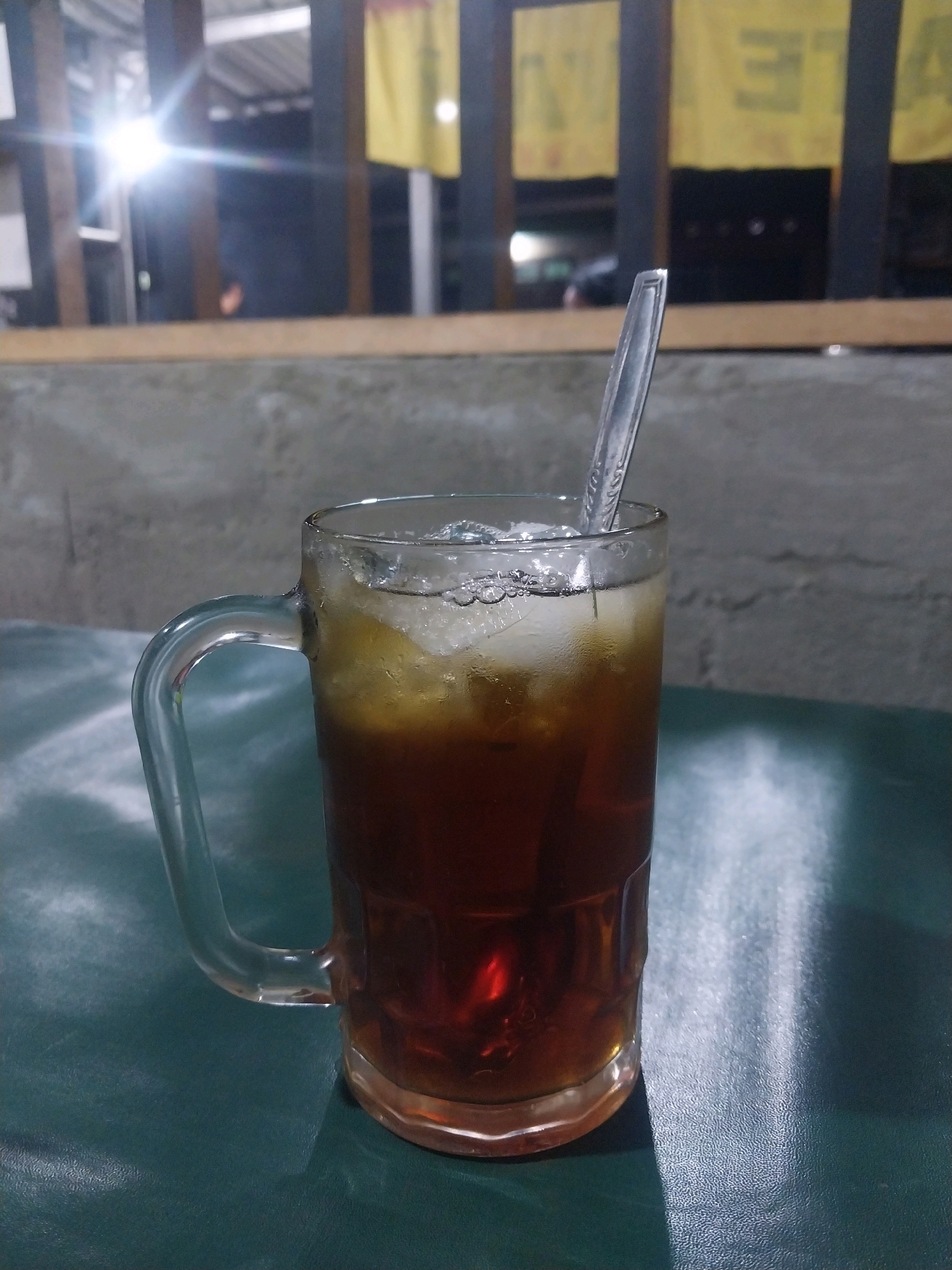 A close-up, eye-level shot captures a tall glass mug filled with iced tea, complete with ice cubes and a clear plastic stirrer. The mug has a sturdy handle and rests on a dark green, textured surface that appears to be a table. The beverage itself is a deep amber color, with condensation visible on the outside of the glass. In the background, a concrete wall with rough texture is visible, and beyond that, a wooden railing separates the foreground from a dimly lit area. Further in the distance, bright yellow banners hang, partially obscured by structural elements. A bright light source is visible in the upper left of the frame, casting lens flares. The overall setting suggests an outdoor or semi-outdoor establishment, possibly a cafe or eatery, located in Trihanggo, Indonesia.