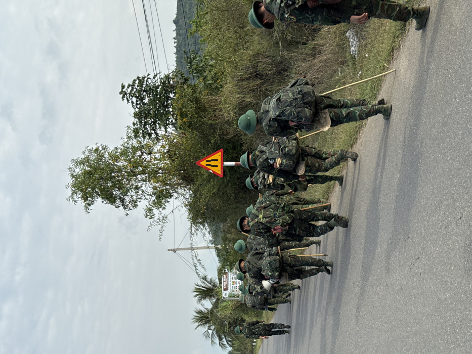 Approximately twelve individuals, identified as soldiers or military trainees, are depicted walking in a column along a paved road in the city of Ninh Hòa, Vietnam. They are viewed from behind, moving away from the camera towards the left of the frame. Each person wears camouflage fatigues, a green pith helmet, and a camouflage backpack. Several individuals have additional gear strapped to their packs, including rolled items and containers, and many carry a long, light-colored wooden staff in their right hand.
The road surface is asphalt, gray in color, and appears to be a two-lane thoroughfare without distinct markings. To the right of the road, dense green and brown vegetation, including bushes and trees, lines the edge. A triangular red and yellow road sign with two vertical black lines (indicating a road narrowing or obstacle) is prominently visible against the vegetation. Power lines are strung between utility poles further in the background on the right. To the left of the road, sparser vegetation and a partially visible large white sign are present. Distant, tree-covered hills or mountains are visible under a cloudy, overcast sky, indicating daylight conditions.