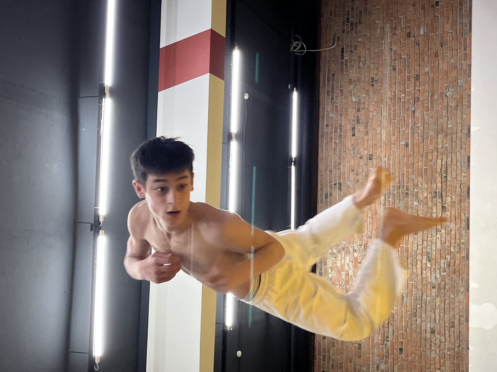 A young, light-skinned man with dark hair is captured mid-air, his body angled towards the right. He is shirtless and wearing white pants. His expression is one of surprise or exertion. The background features an indoor setting with a gray wall on the left, illuminated by vertical light strips. To the right of the man, there is a brick wall and a panel with red, white, and gold coloring. The scene appears to be in an urban environment, consistent with the city of Krasnoyarsk, Russia. There are some cables visible on the right side. The man’s body positioning suggests he is performing a dynamic movement or jump.