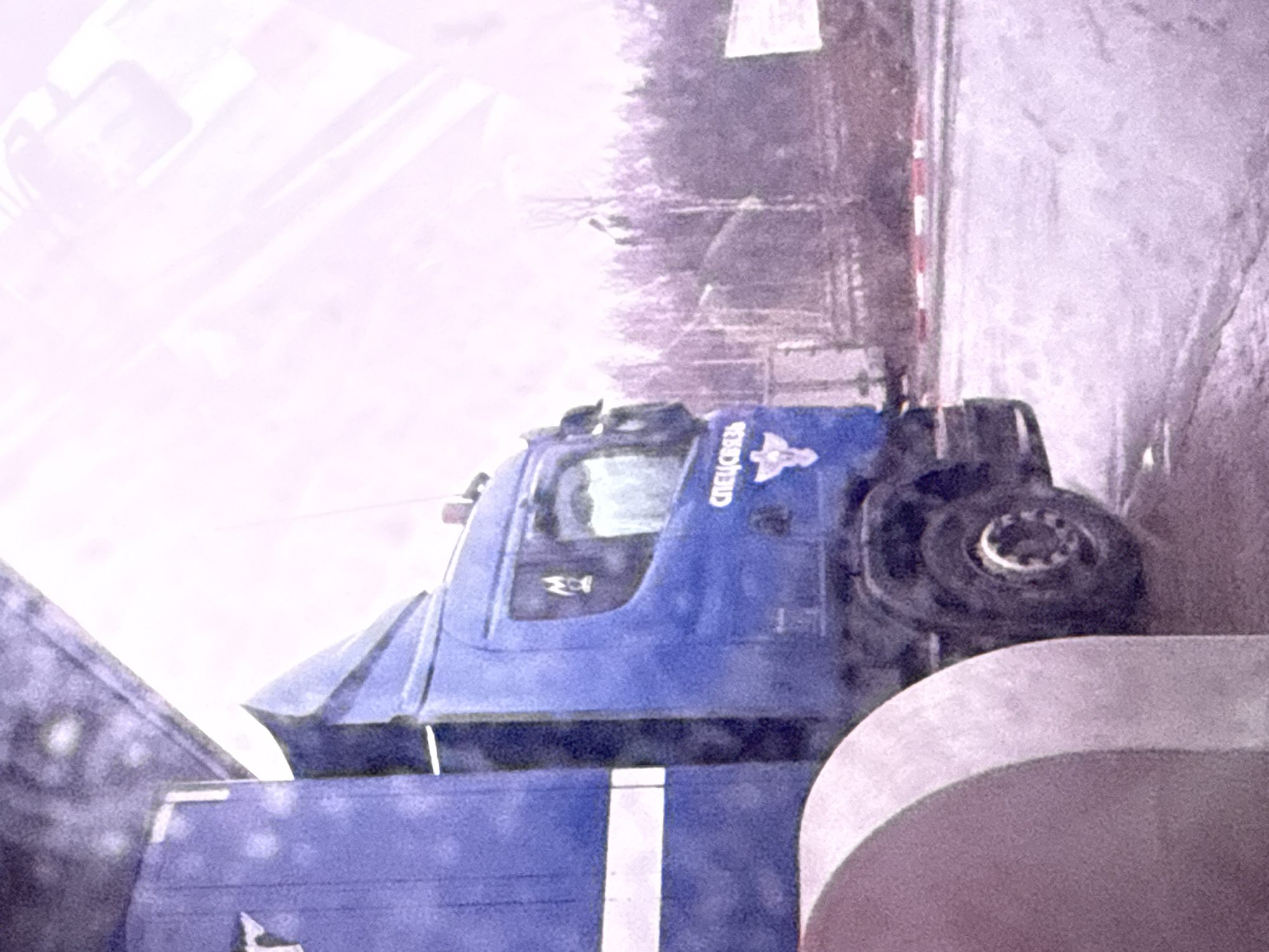 The image displays a large blue heavy-duty truck, possibly a specialized vehicle or a semi-trailer cab, positioned diagonally across the frame. On the side of its cabin, the Cyrillic text "СПЕЦСВЯЗЬ" (SPETSSVYAZ), meaning 'Special Communications,' is clearly visible, accompanied by a stylized eagle or bird emblem. The truck appears to be parked or moving slowly on a paved road or lot.
In the background, a sparse line of trees suggests a rural or semi-urban environment. To the right, a white and red striped barrier post is partially visible. The ground surface exhibits a textured appearance, which could indicate a thin layer of snow, ice, or simply worn asphalt. The overall lighting is very bright and somewhat washed out, casting a high-key, slightly purplish-pink hue over the scene. This bright, diffuse light might suggest an overcast day or highly reflective snow cover.
No people are visible in the scene. A portion of another vehicle or trailer, also in shades of blue and gray with a white stripe, is seen in the lower left and bottom center of the frame, indicating the blue truck might be attached to it or parked adjacently. The setting is Kiselevka, Russia.