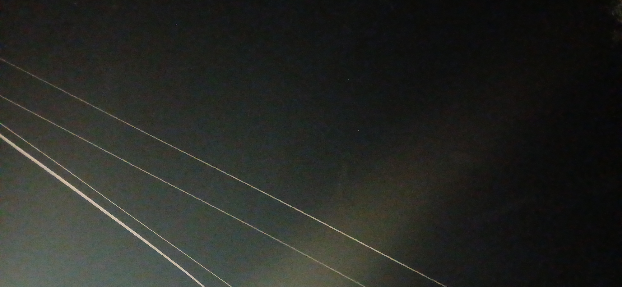 The image is a photograph of power lines against a dark, possibly night-time, sky. Several parallel power lines are visible, running diagonally from the upper left to the lower right of the frame. The lines are thin and light-colored, appearing to be illuminated or reflecting light. The background is predominantly dark green or black, with some subtle variations in tone suggesting a textured surface or a distant, indistinct environment. The overall impression is one of urban infrastructure set against a dark backdrop, possibly taken at dusk or during the night in an open area of Sumy, Ukraine. There are no people or discernible landmarks in the image.