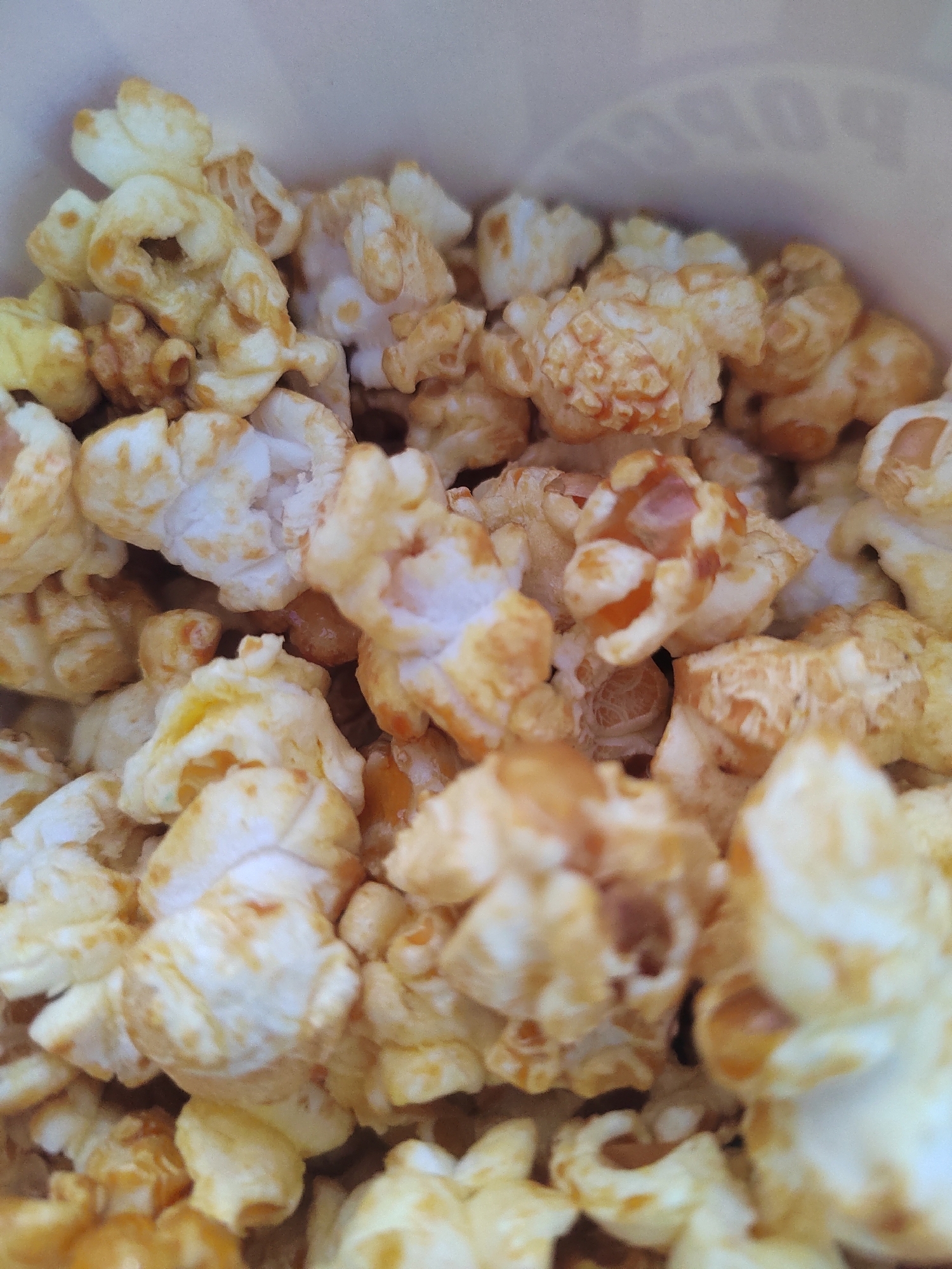 The image is a close-up, top-down shot of caramel popcorn inside a paper container. The popcorn pieces are varied in shape and size, with some appearing fluffy and white, while others are coated in a golden-brown caramel glaze. The caramel gives some pieces a slightly darker, almost burnt appearance in areas. The texture of the popcorn is visible, showing the puffed kernels and the sticky caramel coating. The background is out of focus, but appears to be the interior of the paper container, with a subtle texture and a hint of a logo or text that is not clearly legible. There are no people, animals, or other objects visible in the image. The setting does not provide any information that would allow for the identification of a specific location in Vityazevo, Russia, or any other city.