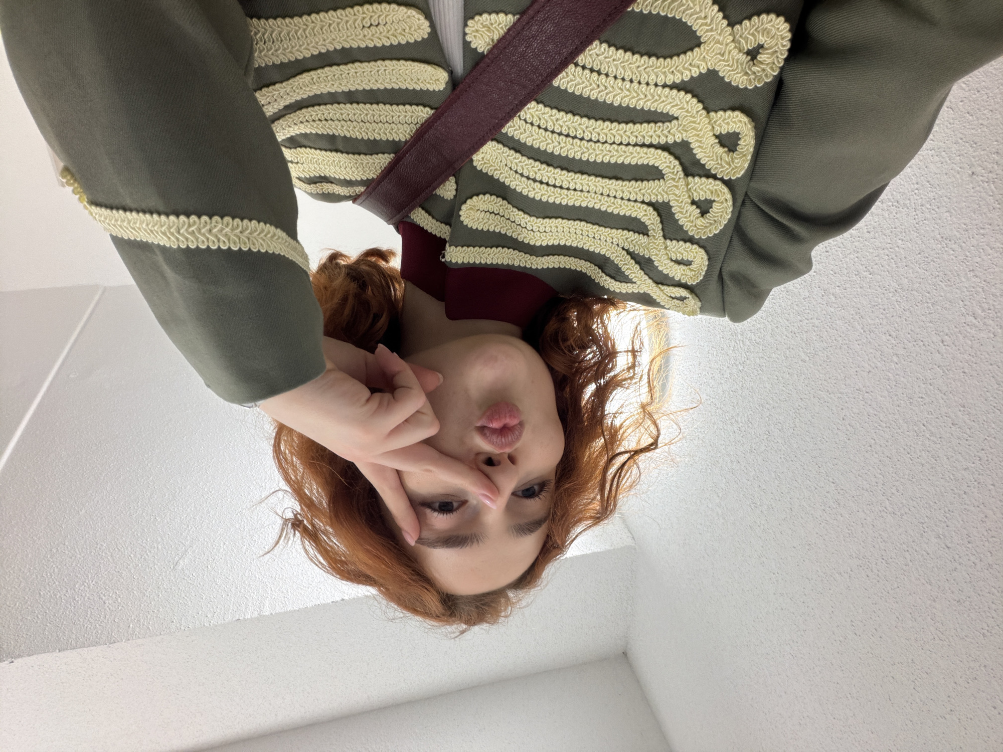 The image shows a person with reddish-brown curly hair looking into a camera, making a kissing face with their fingers in a peace sign gesture near their eye. They are wearing a dark green jacket with ornate cream-colored braided trim on the chest and sleeves, a dark red turtleneck, and a burgundy strap across their chest. The background is a white textured wall and ceiling, possibly an indoor space. There is no indication of the location being the city of Strasbourg, France.