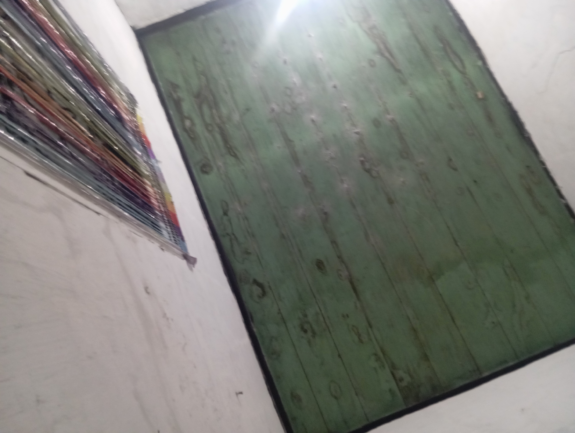 The image depicts a ceiling with a section of wooden planks painted green. Above the wooden planks, a stack of colorful magazines or papers is visible, wrapped in clear plastic. The scene appears to be indoors, with a white wall or ceiling in the background. There is a bright light source coming from the right side of the image, causing a glare on the wooden surface. The environment suggests a storage area or a room where items are kept. The location in Rejosari, Indonesia, implies a tropical or subtropical climate, which might influence the materials and preservation of the objects.