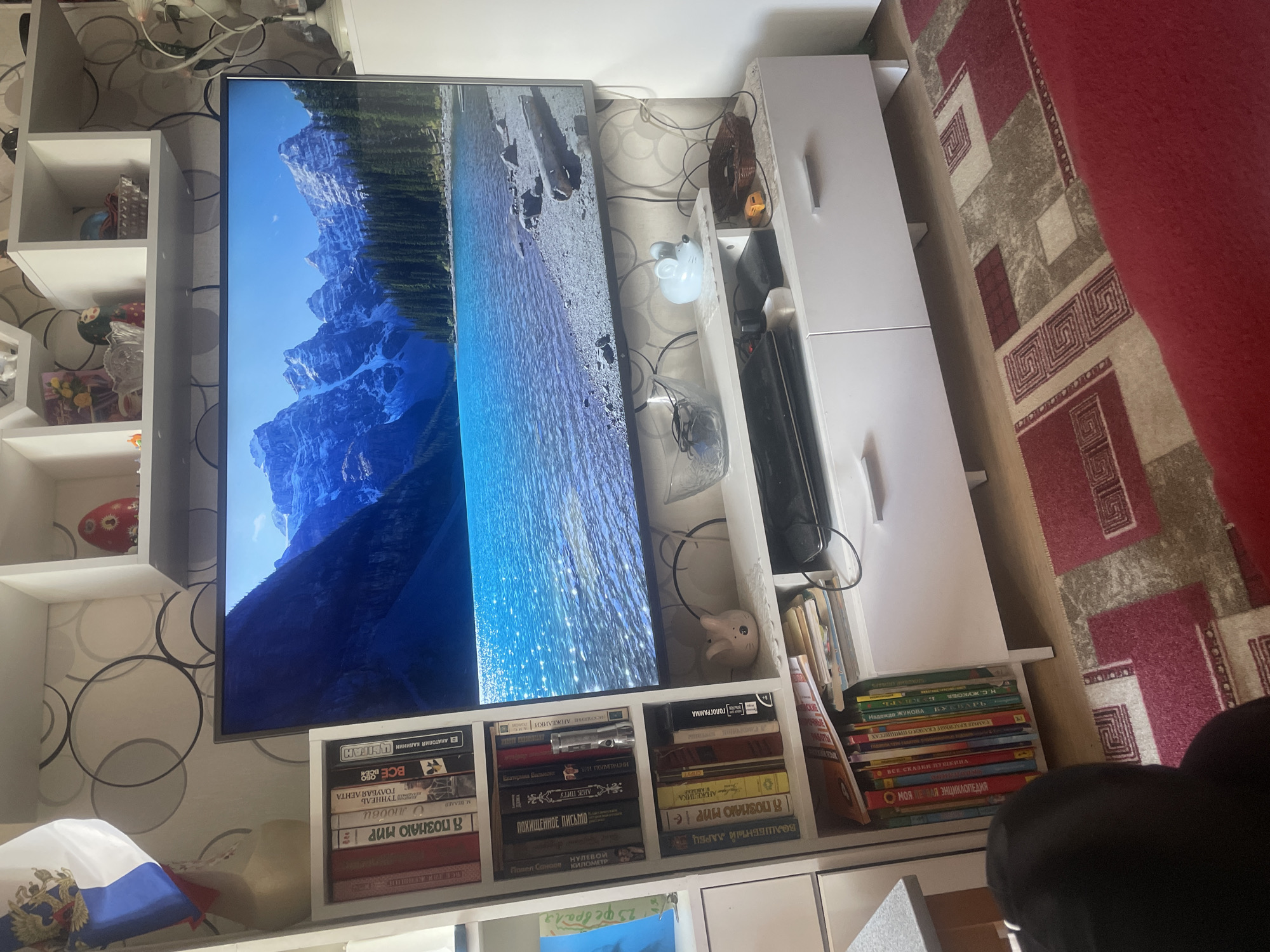 The image depicts an indoor scene, likely a living room, with a large television prominently displaying a landscape of mountains and a body of water. To the left of the television is a white shelving unit filled with books. Above the television, on a white shelf, are various decorative items, including what appears to be a Russian flag. Below the television is a white media console with drawers, and a portion of a red and beige patterned rug is visible on the floor in the foreground. A person's back, wearing a dark top, is partially visible in the lower left corner of the image, suggesting they are facing the television. The overall environment suggests a domestic setting. The provided information indicating the location as Sarov, Russia, is a contextual detail.