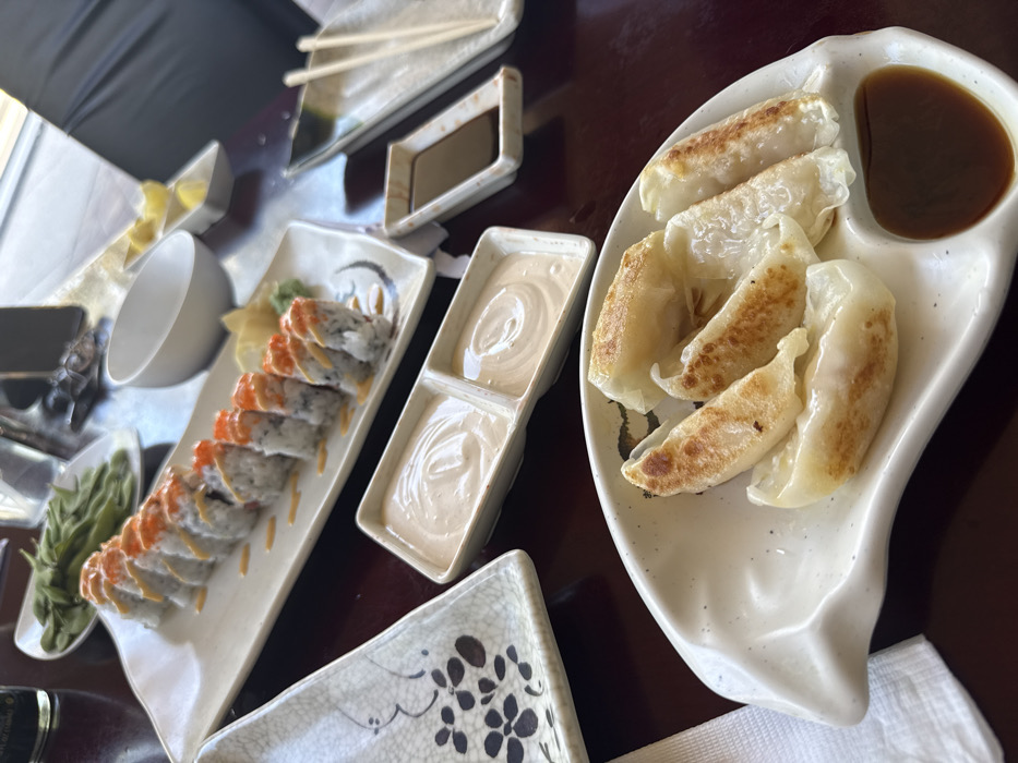 The scene depicts an indoor dining table within an Asian restaurant, consistent with establishments found in Naples, United States. The dark brown table surface holds multiple ceramic dishes. In the foreground, a white, irregularly shaped plate contains five pan-fried dumplings (gyoza), with a separate compartment holding a dark brown dipping sauce. Centrally, a long, rectangular white plate presents ten pieces of a sushi roll, each topped with reddish-orange roe and a light-colored sauce drizzle. Adjacent to the sushi, a two-compartment rectangular dish holds two servings of a creamy, pale sauce. In the background, a white bowl filled with green edamame pods, an empty white bowl, and a small rectangular dish containing lemon wedges are visible. A separate place setting includes a rectangular dish with a dark liquid (likely soy sauce) and a pair of wooden chopsticks. No individuals are present in the frame. The environment suggests natural lighting originating from an unseen window on the right.