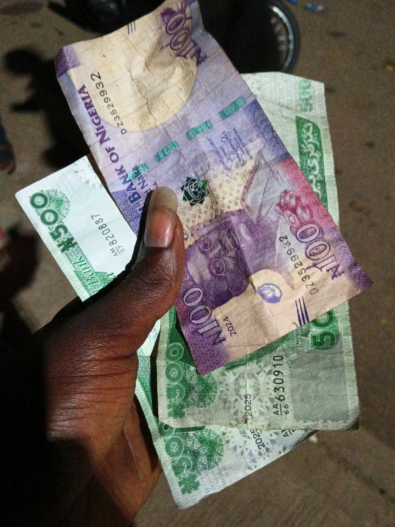 In Bukuru, Nigeria, a hand is shown holding several Nigerian Naira banknotes. The hand, with dark skin and a manicured fingernail, grasps the bills from the bottom. Prominently displayed are a 100 Naira note and a 500 Naira note. The 100 Naira note, predominantly purple and beige, shows the inscription "BANK OF NIGERIA" and a serial number "DZ3529932". The 500 Naira note, with green and white coloring, features the denomination "#500" and serial number "AM 820887". The background suggests an outdoor setting with a dirt or asphalt surface, and the lighting indicates it is likely evening or nighttime.