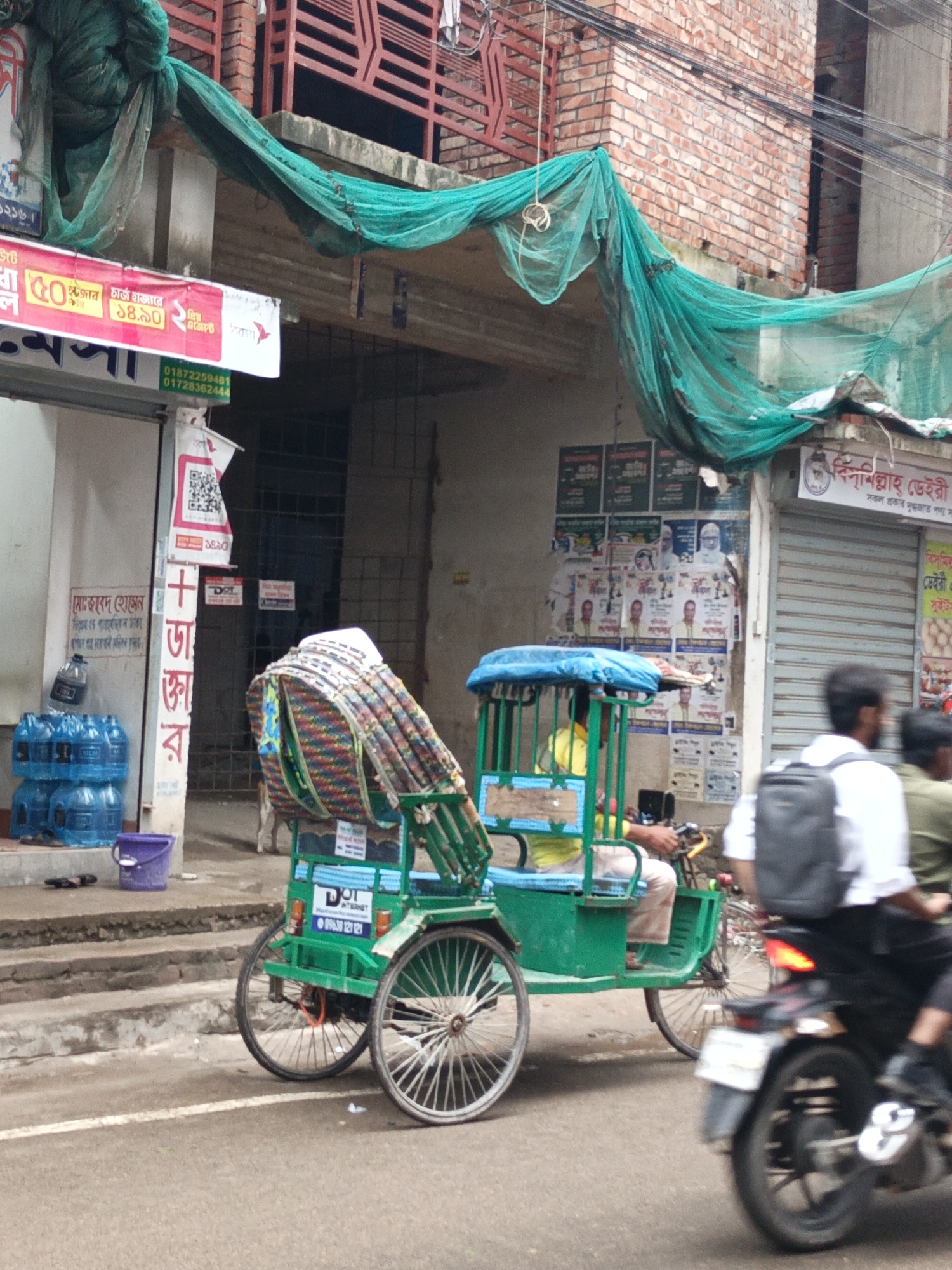 The image captures a vibrant street scene, likely in an urban area of Bangladesh. In the foreground, a green three-wheeled cycle rickshaw is central, with a passenger wearing a white shirt and backpack facing away from the camera. The rickshaw puller is partially visible, wearing a yellow shirt, and the rickshaw has a blue canopy over the passenger seat. To the right, a blurred motorcycle moves past, carrying at least two individuals, with one rider's back and arm visible. The road is asphalt with a white line.
On the left side of the street, there's a sidewalk with blue water bottles stacked near a building entrance, next to a purple bucket. The building itself has a light-colored facade with several posters and advertisements affixed to it. Further back, the building features exposed red brickwork and a metal-grated window or door. Green netting is draped across the top of the street level, connecting to the building. Overhead, electrical wires crisscross.
On the right side of the street, another building with a metal roller shutter is visible, also adorned with numerous posters and advertisements, including many political-style posters featuring faces.
Visible text includes:
- "১২১৬" (1216)
- "চার্জ হাজারে" (Charge per thousand)
- "কুজোর ১৪.৯০২" (Kujor 14.902)
- "মেসা" (Mesa)
- "এজেন্টে" (To agent)
- Phone numbers: "01872259481", "01728362444", "09638 121 121"
- "DOL INTERNET"
- "মোঃজাবেদ হোসেন" (Md. Jabed Hossen)
- "বিস্মিল্লাহ্ ডেইরী" (Bismillah Dairy)
- "সকল প্রকার দুগ্ধজাত পণ্য সা" (All types of dairy products)
- "সুইট" (Sweet)
- A QR code is present on one poster.
The scene depicts a busy daytime environment characteristic of a South Asian city.
