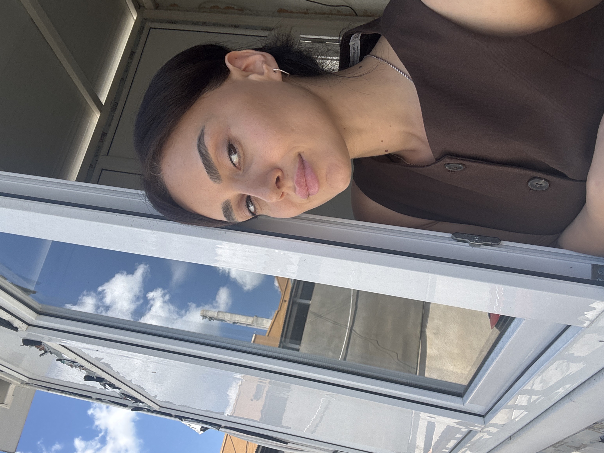 A woman with dark hair and light skin, wearing a brown sleeveless top and a silver necklace, is leaning out of a window. Her expression is a slight smile. The window frame is white, and the glass reflects a blue sky with clouds and some buildings. The environment appears to be outdoors in a city, possibly Tbilisi, Georgia, given the architectural style of the reflected buildings. The overall lighting suggests a daytime scene.