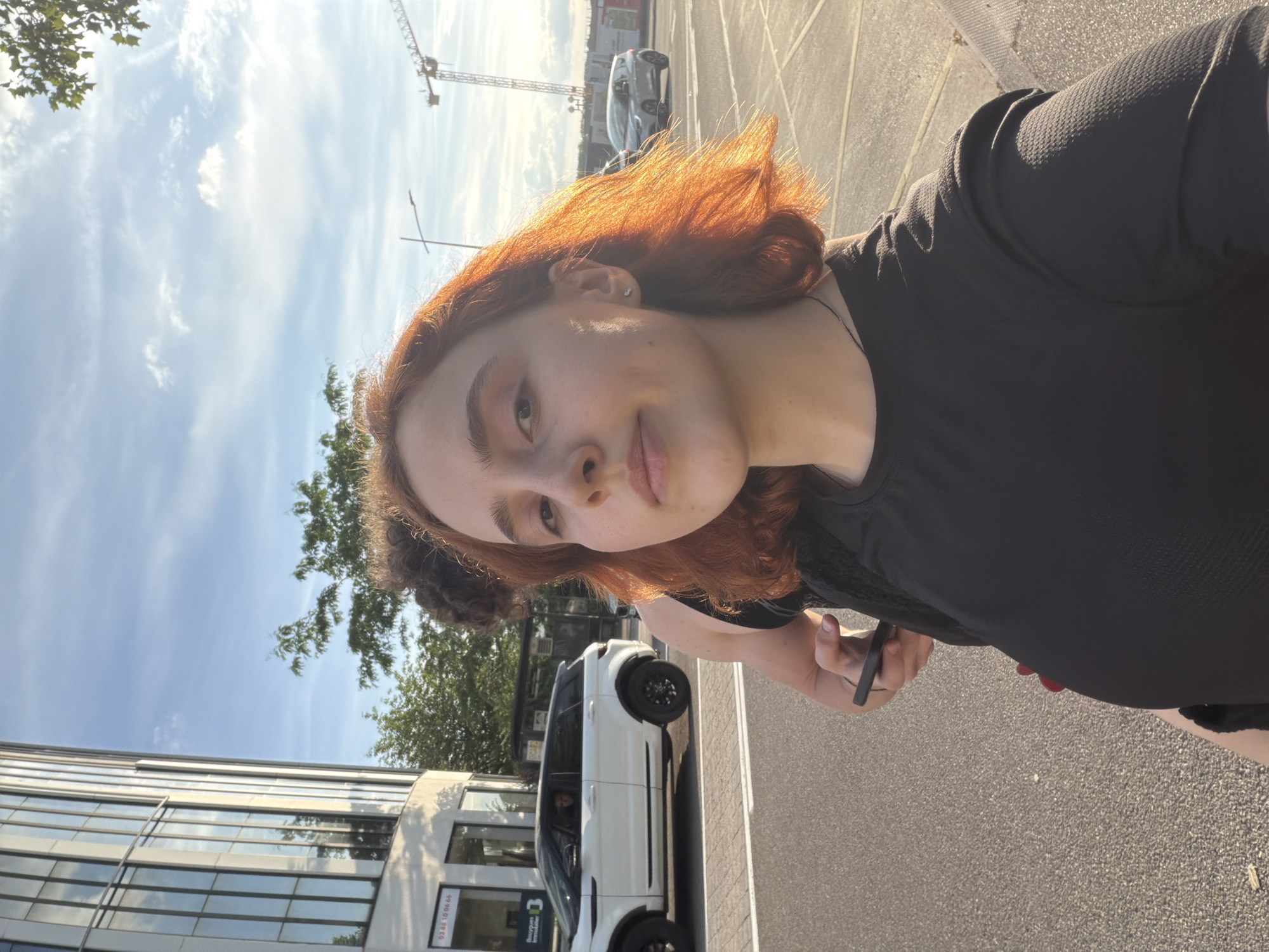 A young woman with reddish-brown hair and a slight smile is captured in a selfie taken outdoors on a bright, sunny day. She is wearing a black textured top and has a stud earring in her left ear. Her right hand is visible, holding a smartphone. A second person with dark hair is partially visible and out of focus behind her.
The setting is an urban environment, likely a street or sidewalk. The sky is clear blue with scattered white clouds, and bright sunlight illuminates the scene, casting strong highlights, particularly on her hair. To the left, a modern building with reflective glass panels is prominent. Behind her, a white SUV (possibly a Range Rover Evoque) is parked on an asphalt street with white markings. Further in the background, another car and a large construction crane are visible against the sky, indicating ongoing development.
On a sign attached to the building behind the white SUV, French text is visible: "03 48 10 06 46" and "BOURGES Immobilier" (meaning real estate). These details confirm the location context as France, consistent with Strasbourg. The overall mood is casual and pleasant on a clear day.