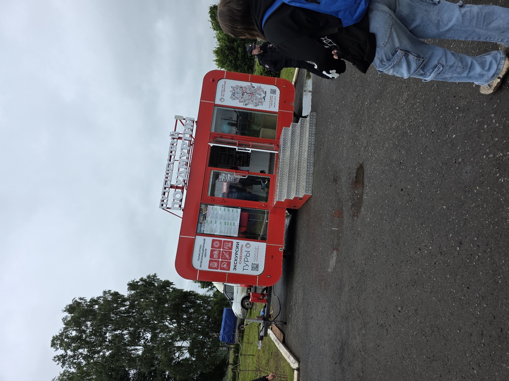 The image shows a red tourist information center trailer labeled "ПУТЕШЕСТВУЙ ПО УДМУРТИИ" (Travel around Udmurtia) situated in the village of Starye Bygi, Russia. The trailer is parked on an asphalt surface, possibly a roadside or parking area, with puddles of water present. A white van and a blue-covered trailer are visible in the background, along with trees and a cloudy sky.
In the foreground, a person wearing a black hoodie, blue cargo jeans, and a blue backpack is facing away from the camera, towards the tourist information center. To the right of the trailer, a police officer in uniform is standing and looking in the direction of the tourist center. Another person's hand is visible on the left side of the image, near a fence. The scene appears to be an outdoor setting, possibly a tourist stop or a public gathering.