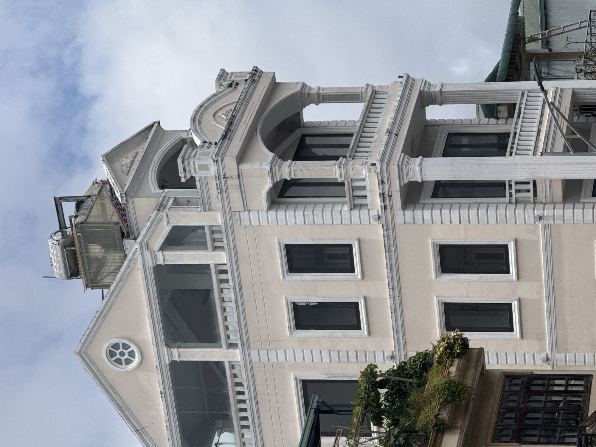 The image shows a multi-story building in Da Lat, Vietnam, with a distinctive architectural style. The building is primarily cream-colored with white decorative elements. The facade features several floors with large, dark-tinted windows. Some floors have balconies with white railings supported by classical-style columns. The top of the building is crowned with a decorative pediment and a circular window with a geometric design. On the roof, a metal water heater and other equipment are visible, along with some of the building's roofing tiles. The sky above is overcast with patches of blue and white clouds. To the left of the main building, a portion of an adjacent building is visible, featuring a darker-colored, multi-paned window and lush green vegetation hanging over a ledge. The overall impression is of a well-maintained, possibly residential or commercial property situated in an urban setting characteristic of Da Lat.