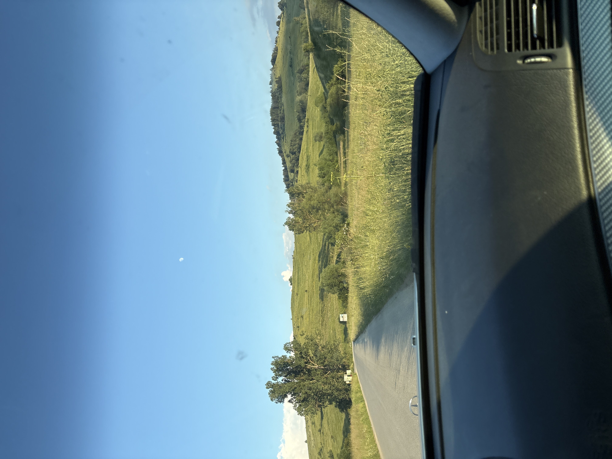 The image shows a view from inside a vehicle looking out towards a rural landscape. The scene is dominated by rolling green hills covered in grass and trees, with a road running through them. The sky is clear and blue, with a small white object, possibly a distant cloud or the moon, visible in the upper left portion of the frame.
On the right side of the image, the interior of the car is visible, including a dashboard with an air vent and a textured panel. The perspective suggests the photograph was taken while the car was in motion, looking forward and slightly to the side.
The time of day appears to be late afternoon or early evening, judging by the warm sunlight illuminating the hills and casting long shadows. The weather seems fair and pleasant. The lush greenery of the hills and the presence of a road indicate a rural or semi-rural setting, consistent with the provided location context of Koprivshtitsa, Bulgaria. There are no people visible in the image. There is no visible text in the image.