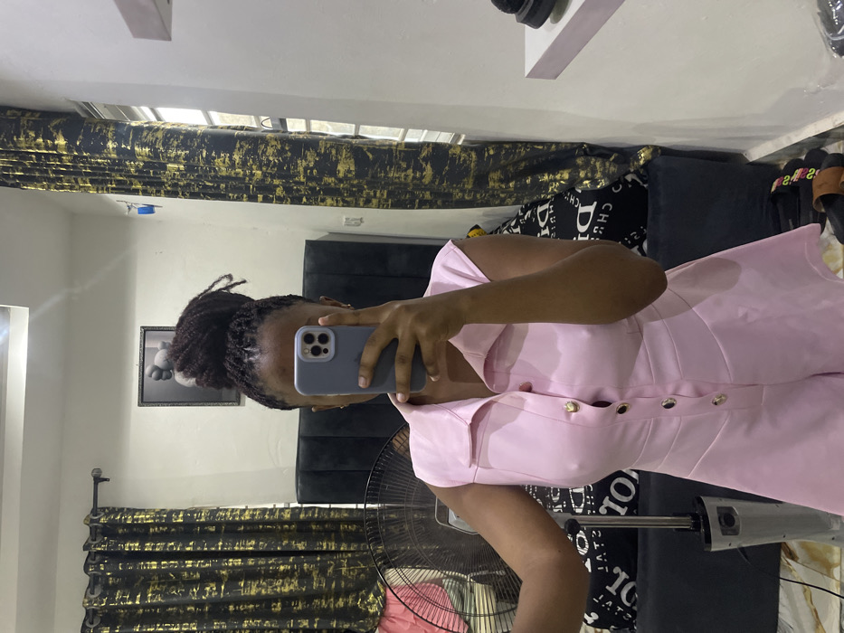 The image displays a female individual taking a mirror selfie inside what appears to be a bedroom. Her face is obscured by a light blue/grey smartphone held at eye level. She possesses dark dreadlocks styled in a high bun. She wears a light pink, sleeveless romper featuring a collar and a front placket with gold-colored buttons.
The room, reflected in the mirror, has light-colored walls. A dark upholstered headboard of a bed is visible behind the individual, with the bed covered by black and white patterned bedding. Above the headboard hangs a framed artwork depicting a stylized grey figure. Dark curtains with abstract gold patterns are drawn across windows on both the left and right sides of the reflection. A metallic pedestal fan stands on the floor to the left of the individual. On the floor in the bottom right, a pair of dark slip-on sandals are visible. A white wall-mounted shelf on the right holds a dark shoe. A small blue object is taped to the wall near the window on the right. This scene is situated within a private residence in Okpanam, Nigeria.