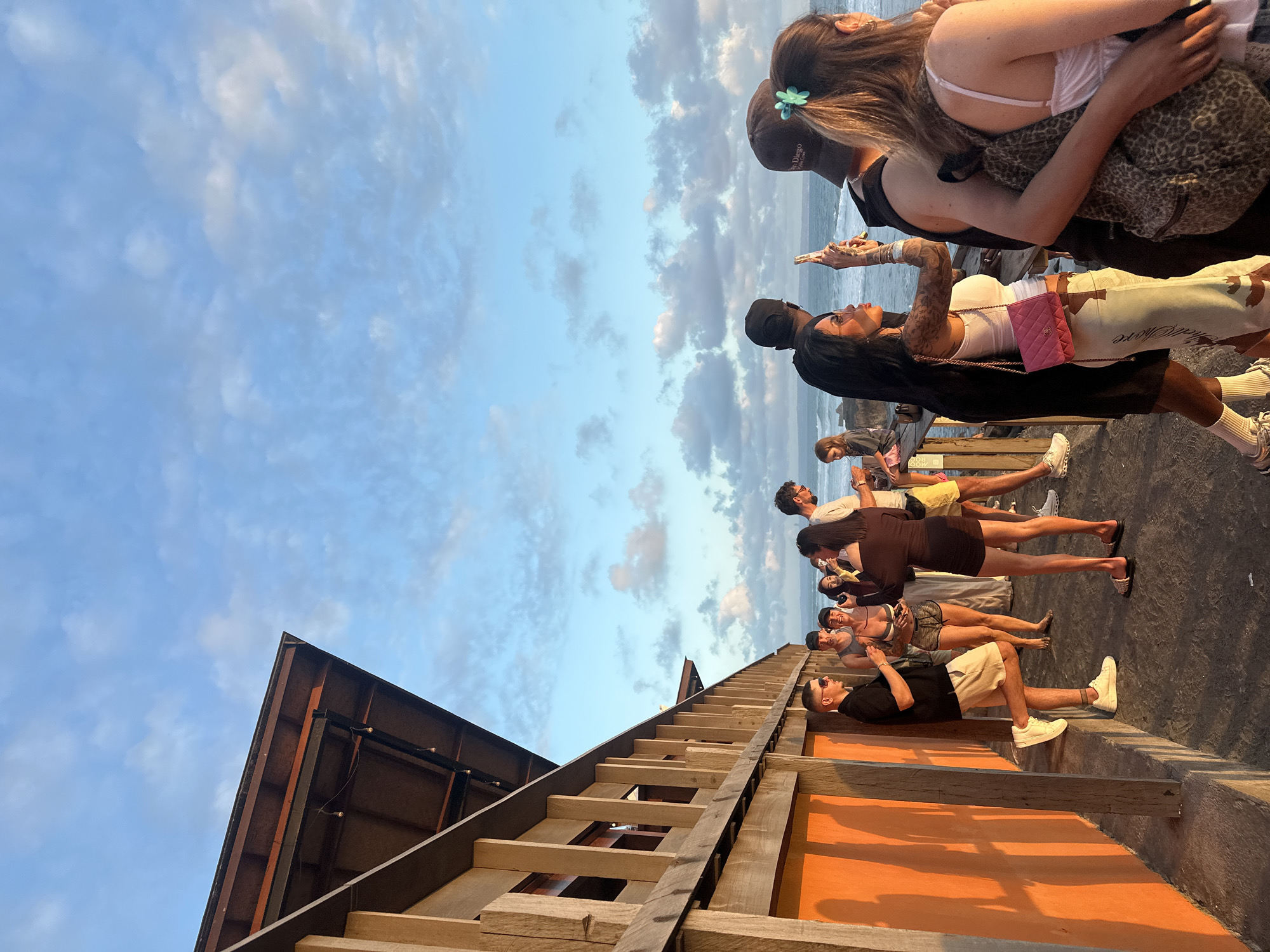 In North Kuta, Indonesia, a group of people are gathered on a wooden deck overlooking the ocean at sunset. The wooden structure, possibly a restaurant or bar, features orange-painted walls and a sloped roof with exposed beams. The sky is a clear blue with scattered clouds, casting warm light on the scene.
Several individuals are visible, some standing and conversing, while others are taking photos or looking out at the sea. One man in the foreground is wearing a black polo shirt and light-colored shorts, with one leg propped on a ledge. To his right, a woman in a black dress stands looking towards the ocean. Further back, a woman with long dark hair is taking a photo with her phone, wearing a white top and a pink quilted purse. Another woman with blonde hair and a green clip is also present.
The environment appears to be a coastal area with waves visible in the background. The overall atmosphere suggests a casual gathering or social event during golden hour.