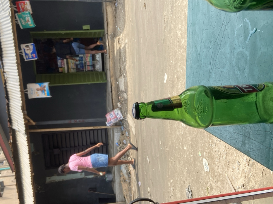 The image depicts a daytime street scene in Alaiyabiagba, Nigeria, viewed from a low perspective. In the immediate foreground, a partially full green glass bottle with a black cap rests on a teal-colored table surface, exhibiting faint liquid spills.
Across an unpaved, debris-strewn road in the midground, two female individuals are observed. One subject, wearing a pink short-sleeved top, a light blue denim skirt, and dark sandals, walks from left to right, carrying a small, light-colored object in her left hand. The second subject, seen from behind in a dark top and light blue shorts, is positioned in the threshold of a small commercial establishment, appearing to enter or exit.
This establishment, situated in the background, features a dark grey facade and open bright green double doors revealing shelves stocked with various products. A corrugated metal roof extends over the entrance, from which multiple advertising bags, including one prominent white bag depicting a baby, are suspended. To the left of this shop, another dark-colored building is visible with a closed, corrugated metal gate. The environment suggests a local commercial area under natural daylight.