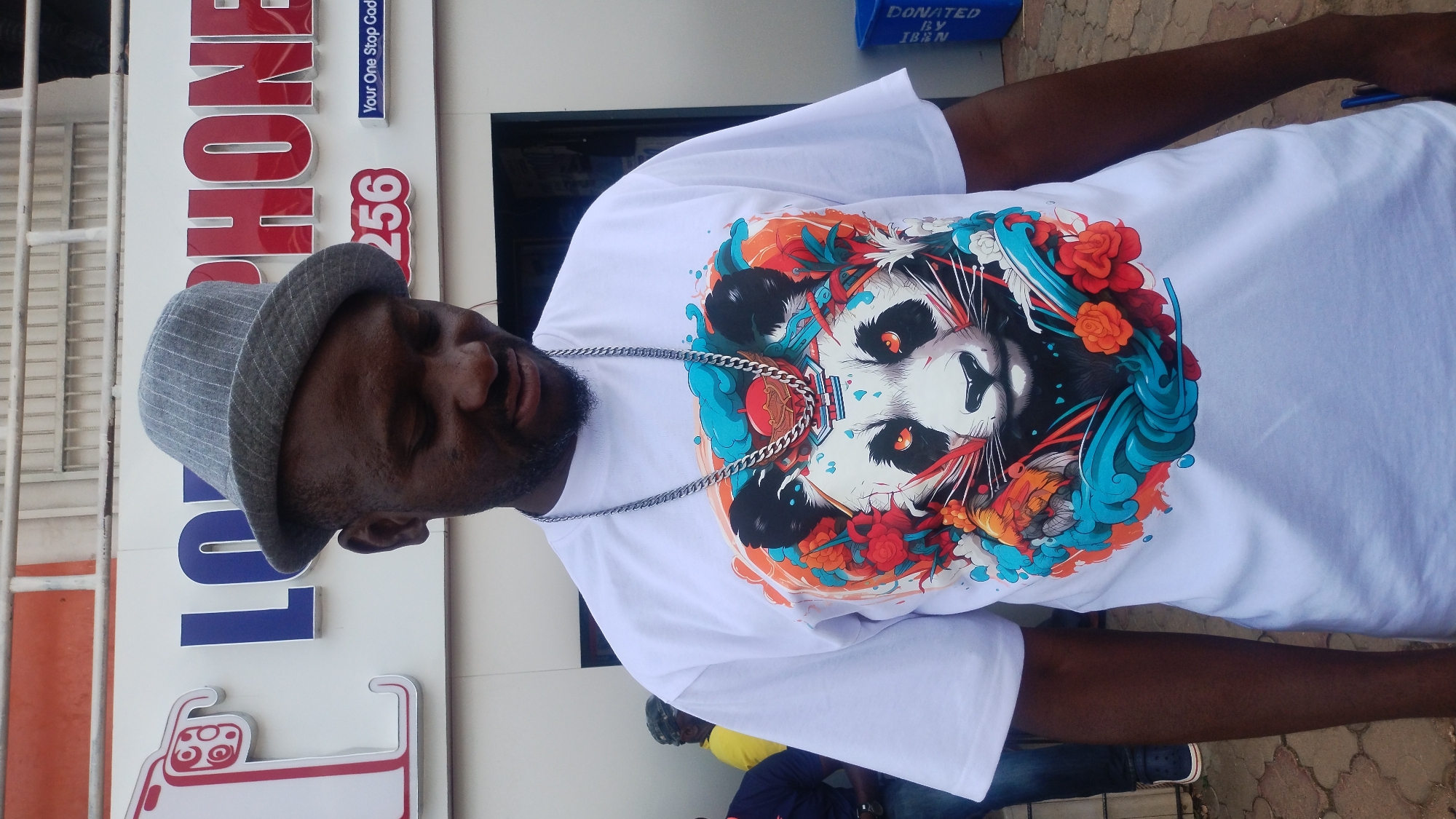 This media file shows a man wearing a white t-shirt with a graphic of a panda and colorful floral elements. He is also wearing a grey, pinstriped hat and a silver chain. The man is standing in front of a building with signs on it. The signs are in red and blue lettering, with one sign saying "PHONE" and another with the number "256". There is also a smaller blue sign that says "Your One Stop Shop". In the background, there are other people, suggesting a public setting. A blue bin with white lettering indicating "DONATED BY IBEN" is visible. The scene appears to be outdoors during the daytime, with a bright, slightly overcast sky, suggesting it could be late morning or afternoon. The overall mood seems casual. The location context provided is Jos, Nigeria.