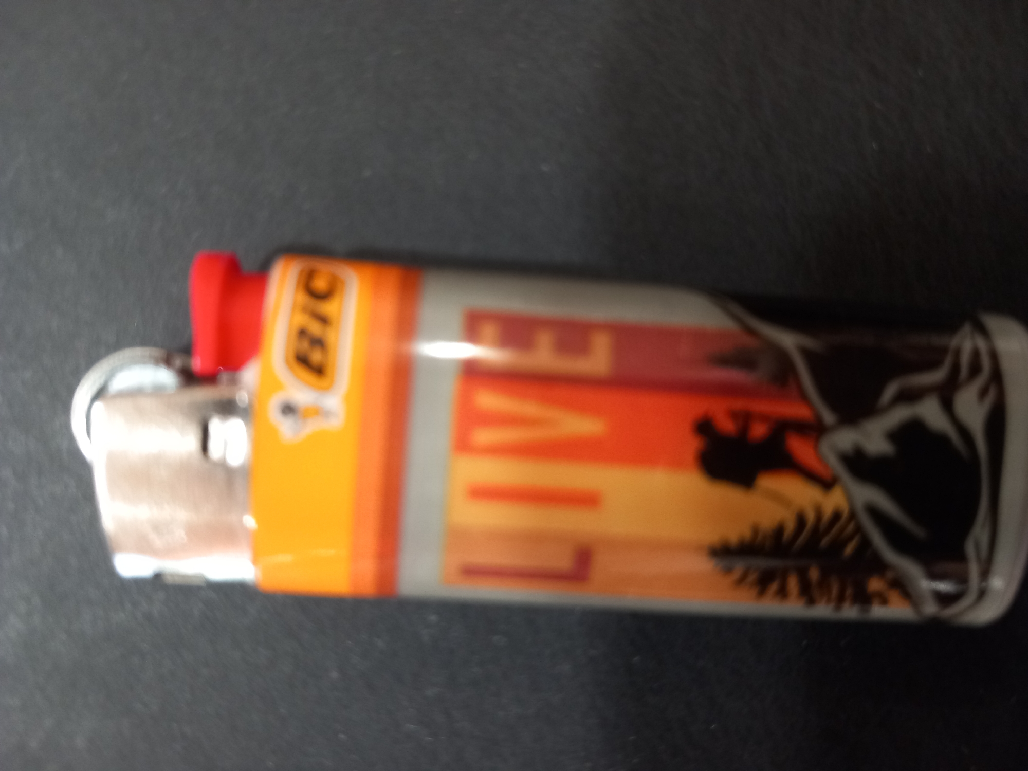 The image shows a close-up, slightly blurry, vertical shot of a BIC lighter on a dark, textured surface. The lighter has an orange top with the BIC logo printed in yellow and black. Below the orange section, there's a clear plastic tube with the word "LIVE" printed vertically in large, bold letters, colored in shades of red, orange, and yellow. The bottom portion of the lighter features a silhouette design of a hiker with a backpack and trekking poles against a sunset or sunrise landscape with mountains and pine trees. The lighting is dim, and the focus is on the lighter. There is no indication of a specific location within a city.