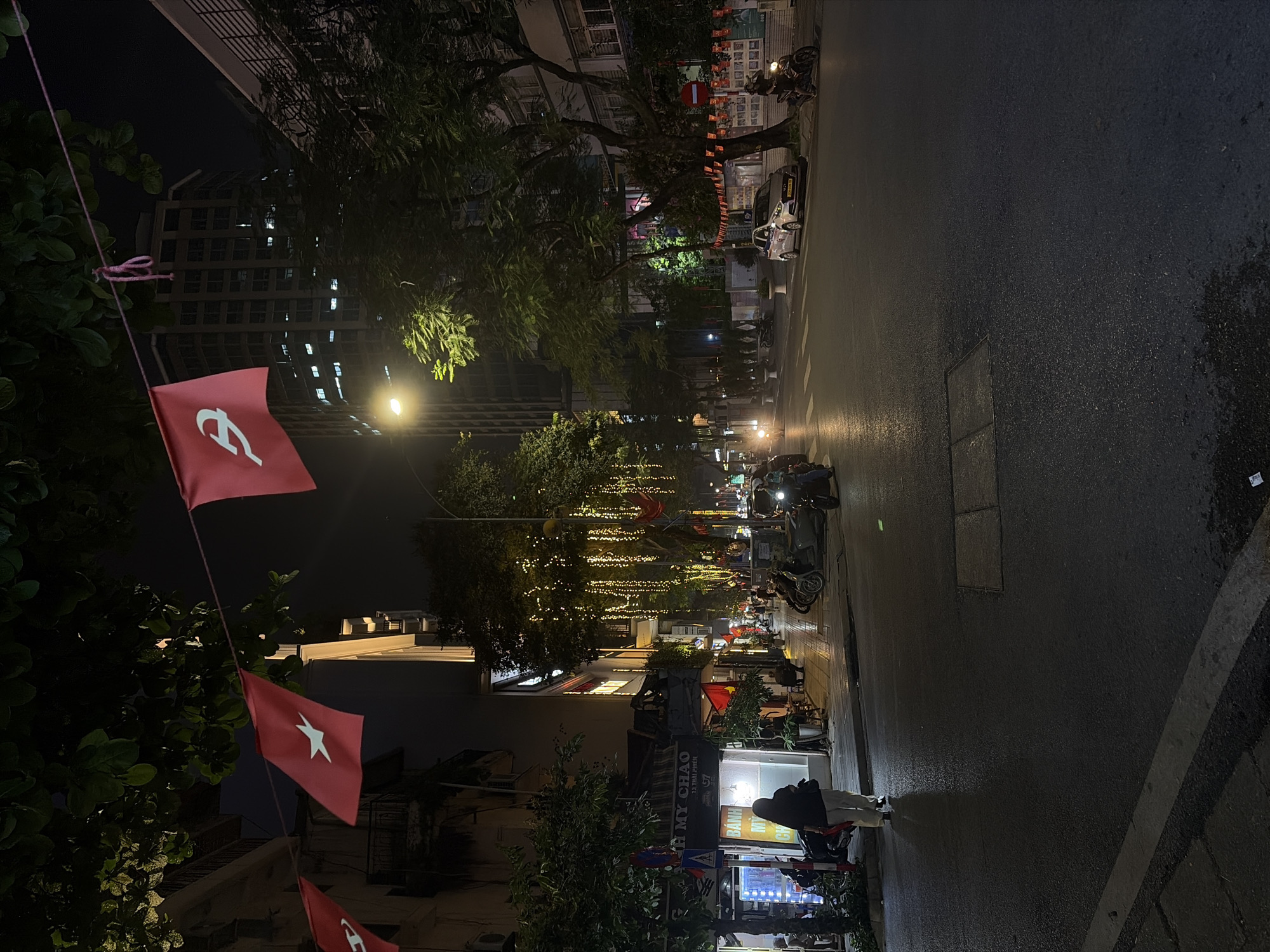 The image depicts a street scene at night in Hanoi, Vietnam, likely in an urban commercial or residential area. Red flags with white star symbols are strung across the street, hanging from trees and poles, suggesting a celebratory or nationalistic occasion. String lights are also visible, illuminating the trees and storefronts. The street is wet, reflecting the artificial lights from buildings and street lamps. Several motorcycles are parked along the sidewalk and on the street. A few people are walking on the sidewalk, and one individual is seen walking away from the camera in the foreground. Buildings line the street, with some displaying illuminated signs. The overall atmosphere is one of a bustling urban environment at night, decorated for an event.