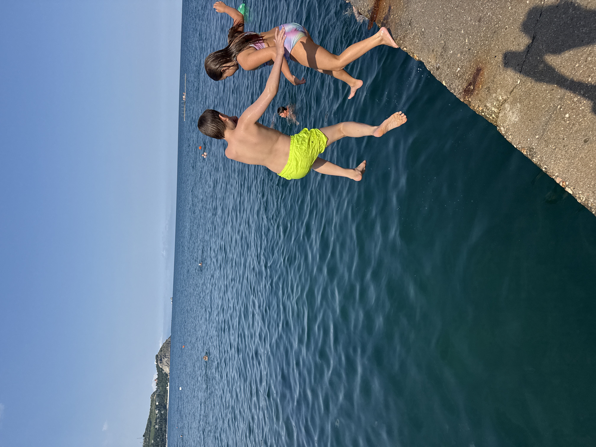 On a bright, sunny day, two children, a boy and a girl, are captured mid-jump as they leap off a concrete pier into the clear blue sea. The boy, wearing bright yellow swim trunks, is to the left and slightly ahead of the girl. The girl, in a multi-colored swimsuit, is just behind him, her body angled as she leaps. Both children are barefoot. In the background, the calm sea stretches out to the horizon under a vast, cloudless blue sky. The coastline of Partenit, Ukraine, is visible in the distance, with green hills and some buildings dotting the landscape. Other people can be seen swimming and enjoying the water further out. A shadow of an adult is cast on the pier, suggesting their presence.