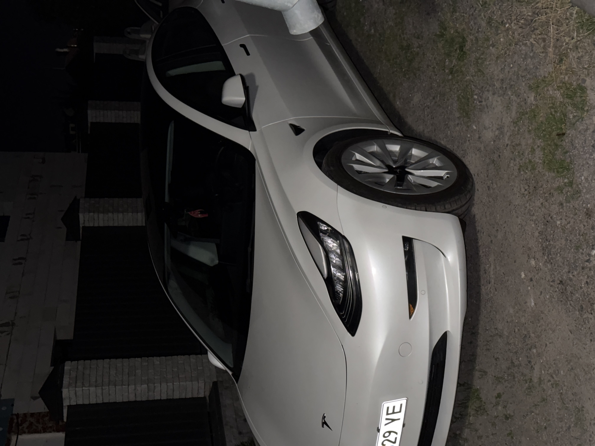 In Bila Tserkva, Ukraine, a white Tesla Model 3 is parked on a gravel surface at night. The car's headlights are on, illuminating the front of the vehicle and its license plate, which reads "29 YE". The car is positioned in front of a dark fence with brick pillars. The scene is dimly lit, suggesting it is nighttime.