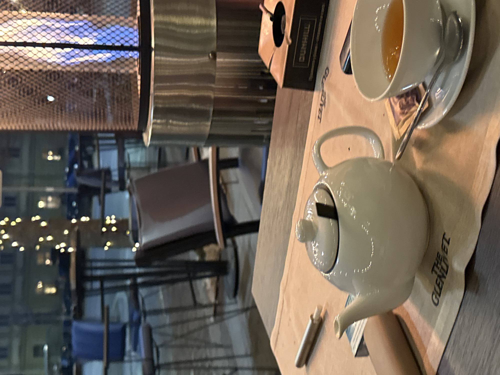 The image depicts an outdoor seating area of a commercial establishment, possibly a cafe or restaurant, in an urban environment in Unknown, Unknown, at night.
In the foreground, a dark wooden table holds a light brown paper placemat branded with "THE GLENLIVET." On the placemat, a white ceramic teapot with a lid and a dark insert is positioned left of center. To its right, a white ceramic teacup filled with an amber liquid (tea) rests on a matching saucer with a silver spoon. A small, light-colored packet lies next to the teacup. Further right, a dark brown and black "DUNHILL" branded ashtray is visible, alongside a dark rectangular object, possibly a lighter or electronic device. To the left of the teapot, a silver cylindrical object, potentially a cigar holder or vape pen, with a white tip, is on the placemat.
In the midground, a stainless steel or metallic outdoor patio heater with a mesh screen displays a visible blue flame. A brown upholstered chair with a dark frame is partially visible, facing away from the camera. In the blurred background, several tall, dark-framed bar tables and stools, some with blue seat cushions, are arranged. A tree trunk wrapped with yellow string lights is prominent. Distant buildings and streetlights are faintly discernible through the general dim ambient light, indicating an evening setting. No individuals are present in the scene.