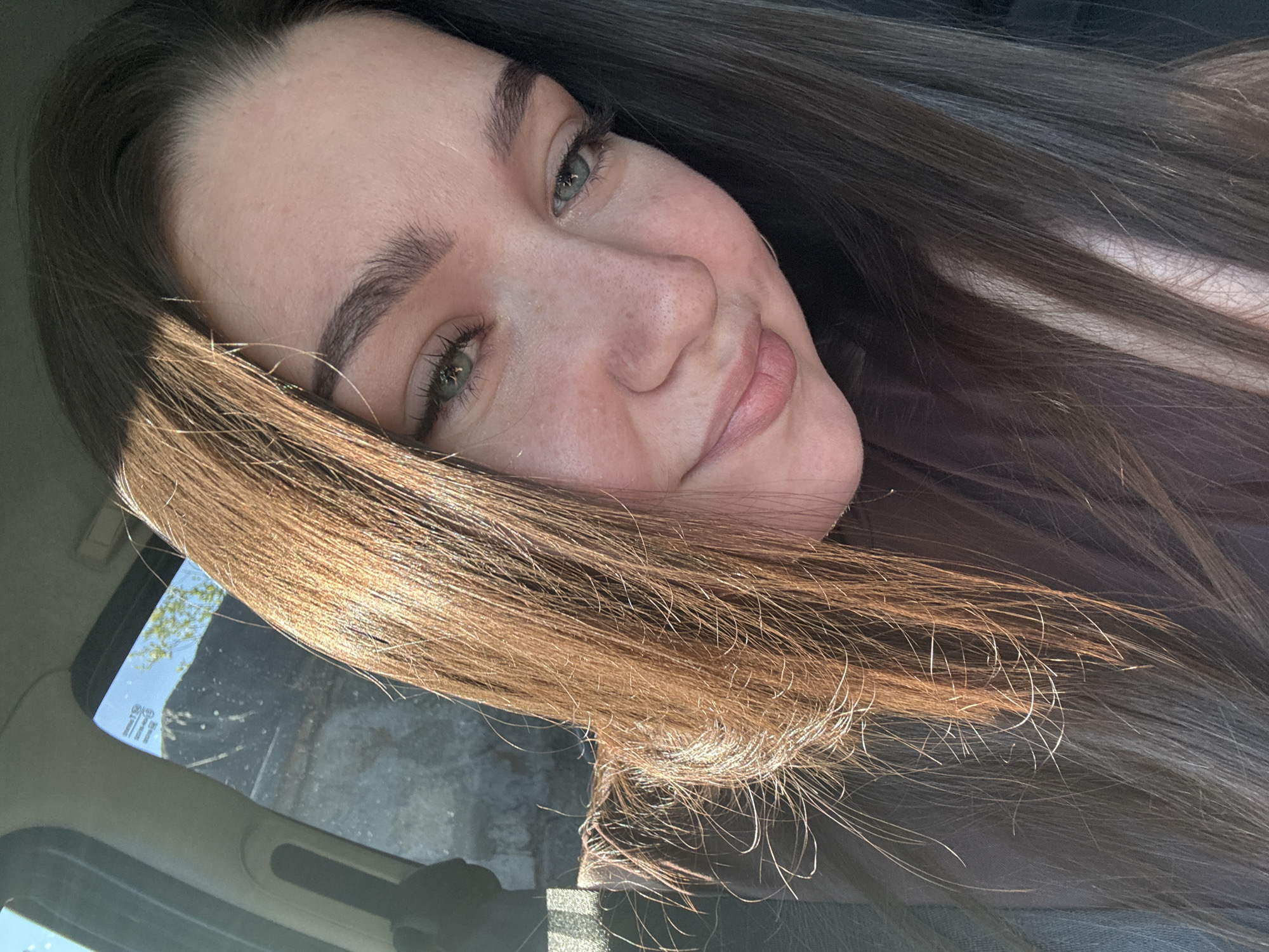 A woman with long brown hair is pictured in a close-up selfie, likely taken inside a vehicle. Part of a car interior is visible in the lower and left portions of the frame. A window can also be seen with an exterior view that includes a tree branch, a building, and a patch of blue sky. The location appears to be Гамово, Russia.