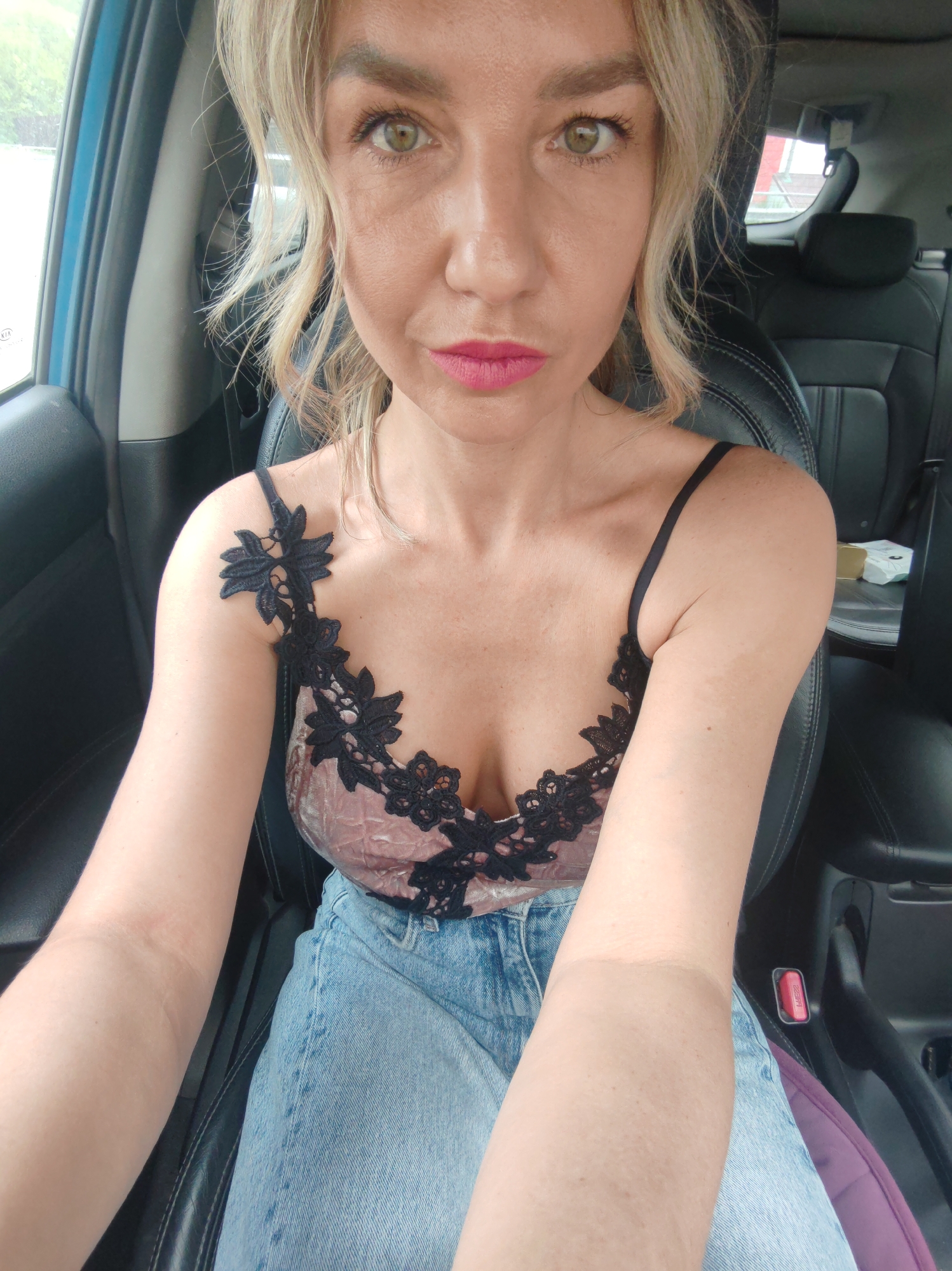 A woman is sitting in the driver's seat of a car, facing the camera. She has blonde hair and is wearing a pink camisole with black lace trim and blue denim jeans. Her eyes are green and her lips are painted pink. The interior of the car is visible, including the black leather seats and the blue door panel. The background is blurred, but suggests an outdoor setting, possibly a street or parking lot in Petropavlovsk-Kamchatsky, Russia. The lighting is natural, indicating it is daytime. The woman appears to be taking a selfie.