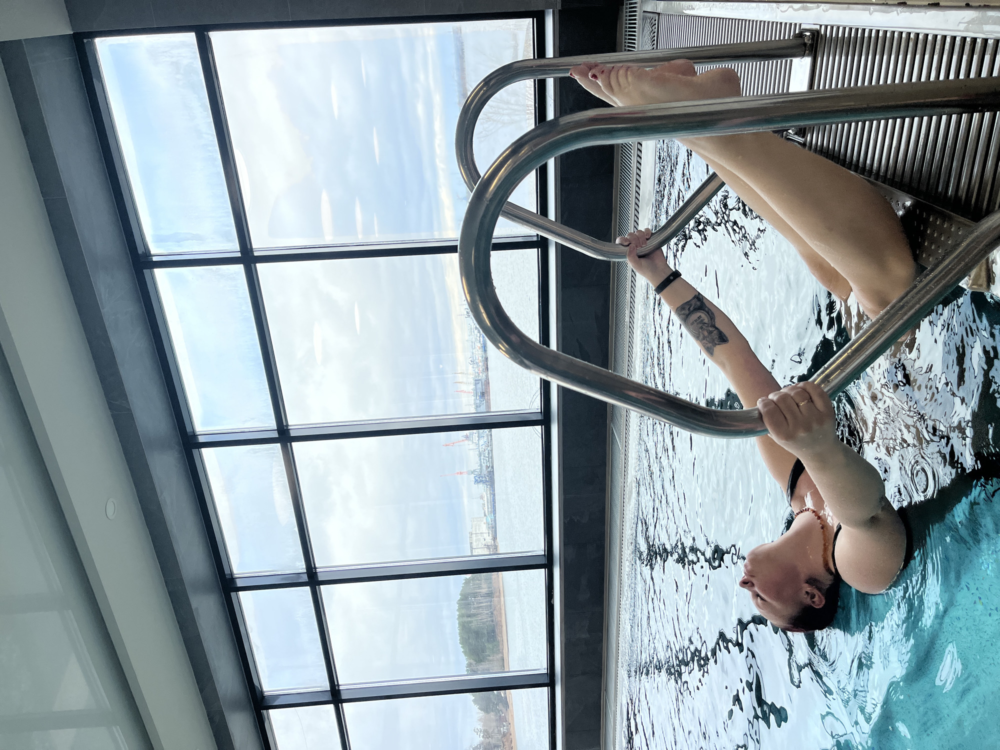 The image captures an indoor swimming pool scene, rotated, showing a person relaxing in the water against a panoramic backdrop of an outdoor landscape seen through large windows.
A person is partially submerged in the light blue pool water, holding onto a stainless steel pool ladder with both hands. Their head and upper torso are in the water, appearing relaxed with eyes closed or looking upwards. Their legs are extended out of the water, resting on a grated section at the pool's edge. A tattoo is visible on their left forearm, and they are wearing a dark swimsuit top and a beaded necklace. The pool water shows ripples and reflections.
The interior features modern design with large, black-framed, floor-to-ceiling windows that offer an expansive view. These windows reveal an outdoor scene under a bright, overcast sky, indicating daytime. A vast body of water, possibly a river or reservoir (consistent with the Kama River in Chaikovsky, Russia), stretches across the mid-ground. The banks or surface of the water show signs of ice or snow, suggesting a winter or early spring setting. In the distance, across the water, industrial elements including cranes and buildings are visible, indicative of a port or urban development. Further along the horizon, a line of trees can be seen.
The atmosphere conveyed is one of calm and relaxation within a modern indoor setting that provides a direct visual connection to the natural and industrial landscape outside. No visible text is present.