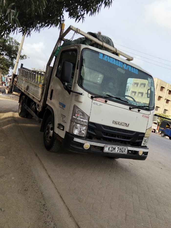 The image features a white Isuzu ELF flatbed truck parked on the unpaved shoulder of a road, captured from a low, front-right angle. The truck is the primary subject, occupying the majority of the frame.
The truck is light-colored, predominantly white, with a dark grille and bumper. The word "ISUZU" is prominently displayed in silver lettering on the front grille. Below the windshield on the passenger side, the model name "ELF" is visible. On the top center of the windshield, blue text reads "M.KORBANI-U/WARE". A small red sticker with "SOYVASA" is located on the upper right side of the windshield. The truck is equipped with a sturdy roof rack, on which some bundled items, possibly tarpaulins or covers, are secured by a wooden frame. The front bumper has two circular yellow auxiliary lights. The license plate is clearly visible on the front bumper, displaying "KDM 760X". The truck's body is a flatbed design with metal side rails, suggesting it's used for transporting goods. On the driver's side door panel, there are several lines of smaller blue text, possibly company information or contact details, but they are too small to be legible.
The setting appears to be an urban or semi-urban area. The truck is parked on a dirt or gravel shoulder, adjacent to a paved road that extends into the background. To the left, a large tree with dense green foliage partially overhangs the scene, providing shade. Behind the tree, a utility pole and faint figures of people are discernible. In the background to the right, a multi-story building with numerous rectangular windows or openings is visible. Further down the paved road, a small blue three-wheeled vehicle, possibly a tuk-tuk, can be seen.
The time of day appears to be daytime, with a bright sky showing some scattered clouds, indicating good visibility. The lighting is even, without harsh shadows, suggesting either late morning or early afternoon. The overall impression is a roadside scene in a populated area. The photo is taken with a slight tilt, making the truck appear angled.
