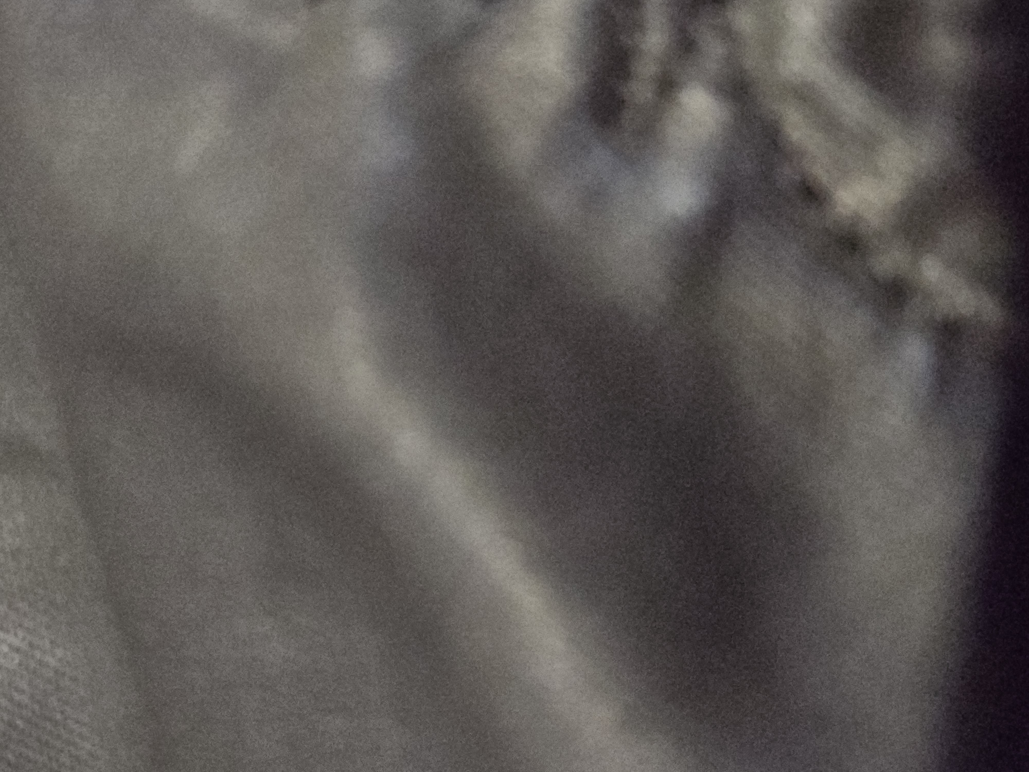 This is a close-up, abstract photograph of a textured, dark-colored fabric, likely silk or a similar material, due to its sheen and the way light reflects off its folds. The fabric appears to be gathered or draped, creating subtle highlights and shadows that give it a dynamic, almost liquid quality. The color palette is muted, consisting of various shades of gray, charcoal, and hints of a very dark, almost black hue. There is a prominent diagonal band of brighter light across the center of the image, suggesting a strong light source reflecting off the smooth surface of the material. The texture of the fabric is visible as a fine grain or weave, adding depth to the otherwise smooth appearance. The overall impression is one of subtle elegance and understated luxury, with the focus solely on the interplay of light, shadow, and texture within the fabric. No people, text, or specific setting cues are discernible. The image is abstract and could be a detail shot of clothing, upholstery, or drapery.