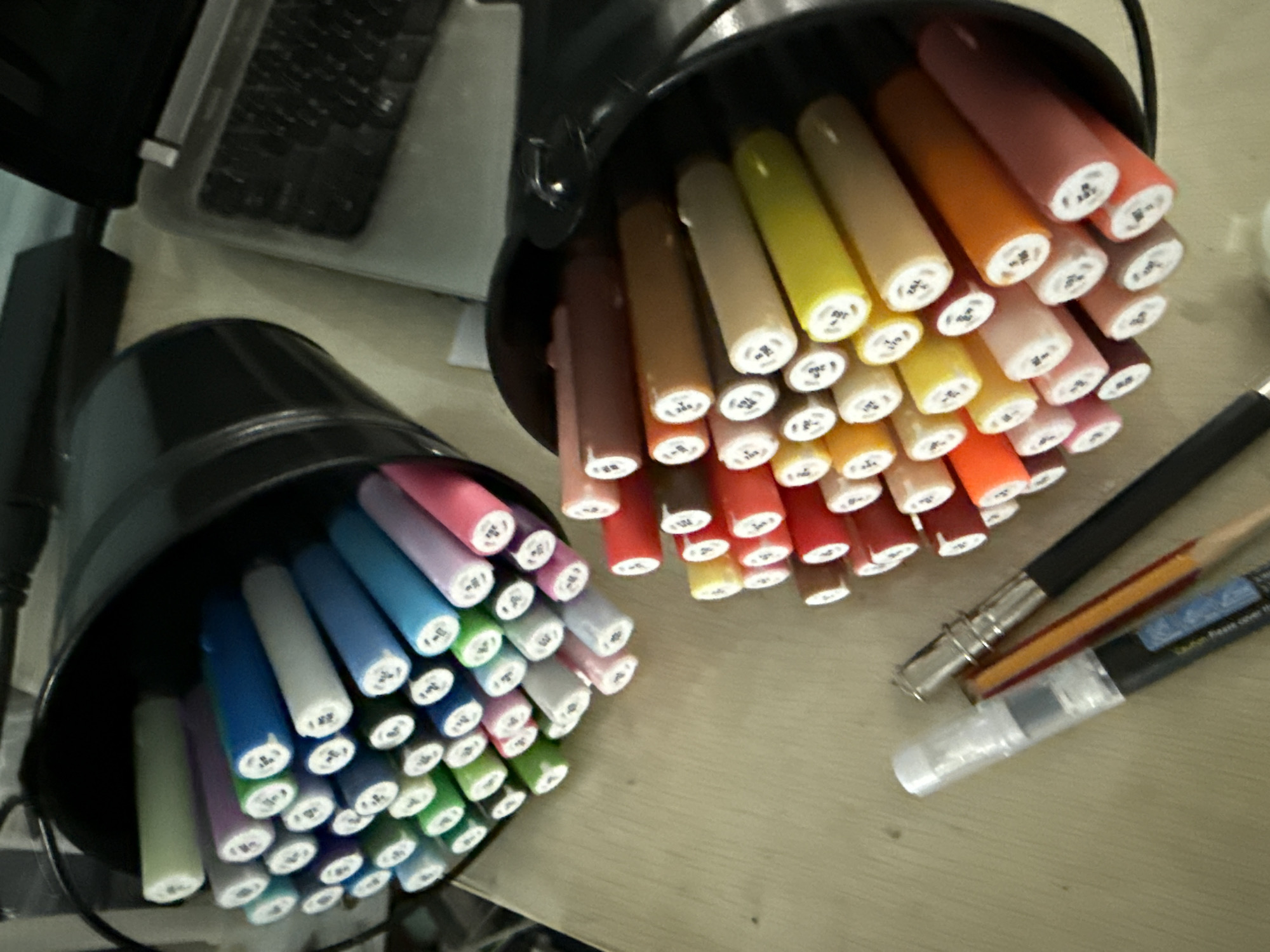 The image shows a close-up, overhead view of several markers in black cylindrical holders, arranged on a light-colored, textured surface that appears to be a desk or table. One holder is in the foreground on the left, filled with markers in various shades of green, blue, purple, and white. The markers are angled upwards and outwards, with their white tips visible.
To the right and slightly behind the first holder is another black cylindrical holder, also filled with markers. These markers are in warmer tones, including shades of orange, red, pink, brown, and yellow. They are also angled upwards and outwards, with their white tips showing. Some of the white tips have small black numbers or symbols printed on them.
In the lower right quadrant of the image, there are several writing instruments lying on the surface. One is a black marker with a silver metal ferrule, a sharpened pencil with an orange body, and a clear plastic barrel with a black cap, which appears to be a water brush pen.
The lighting suggests an indoor setting, possibly during the daytime. There are no people visible in the image. The overall impression is one of an art or craft supplies being organized or prepared for use. The location context of Vladivostok, Russia, does not provide any specific visual cues within the image itself.